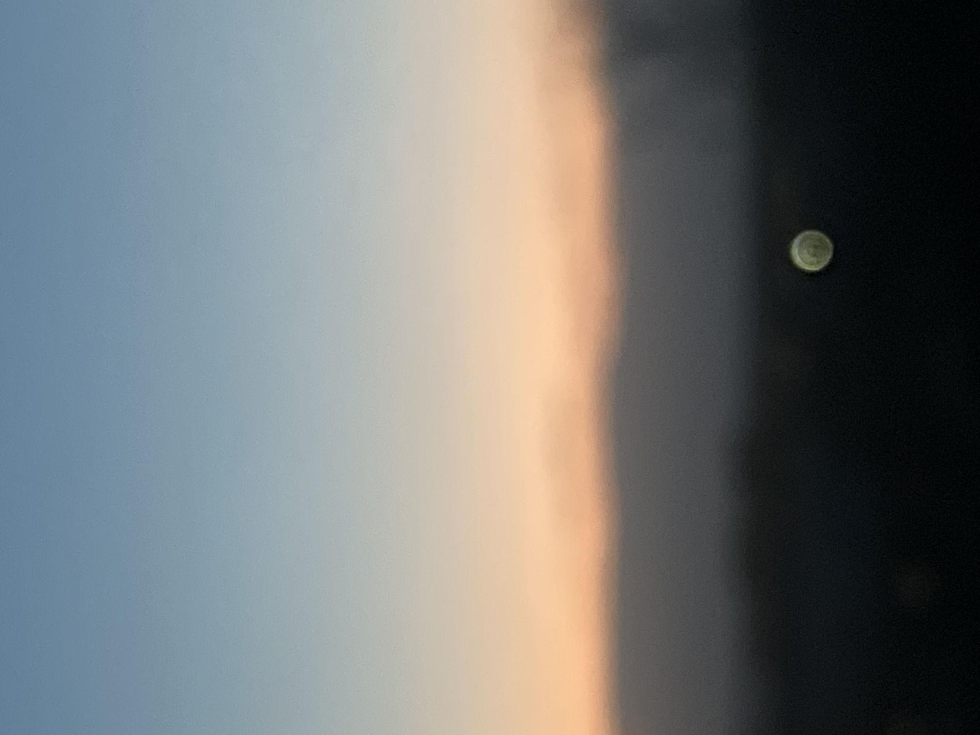 The image shows a sky transitioning from light blue on the left to peach/orange in the center, and then dark gray/black on the right. A circular, greenish shape is visible in the dark area. There are no people or recognizable objects. The location in Votkinsk, Russia is not discernable from the provided image.