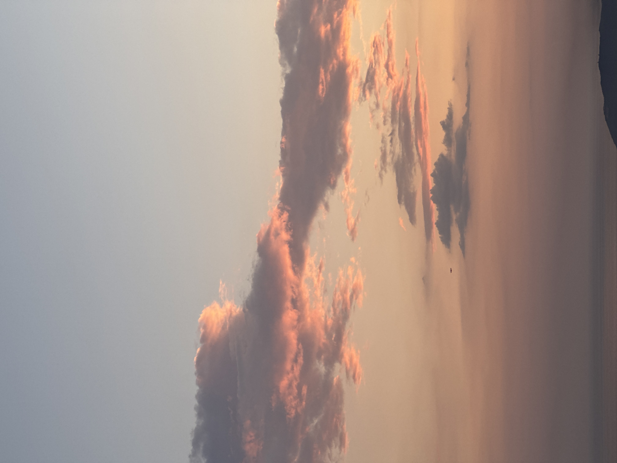 The image captures a twilight scene, likely at dusk, near Sveti Stefan, Montenegro, as indicated by the presence of what seems to be a coastal or mountainous terrain faintly visible on the right edge. The sky occupies most of the frame. The sky transitions from a pale blue on the left side to shades of orange and pink towards the right, suggesting a sunset or sunrise. Various cloud formations are present, some illuminated by the setting or rising sun, casting a reddish-pink glow. A small dark object, potentially a bird or a small aircraft, can be discerned in the middle-right of the frame.