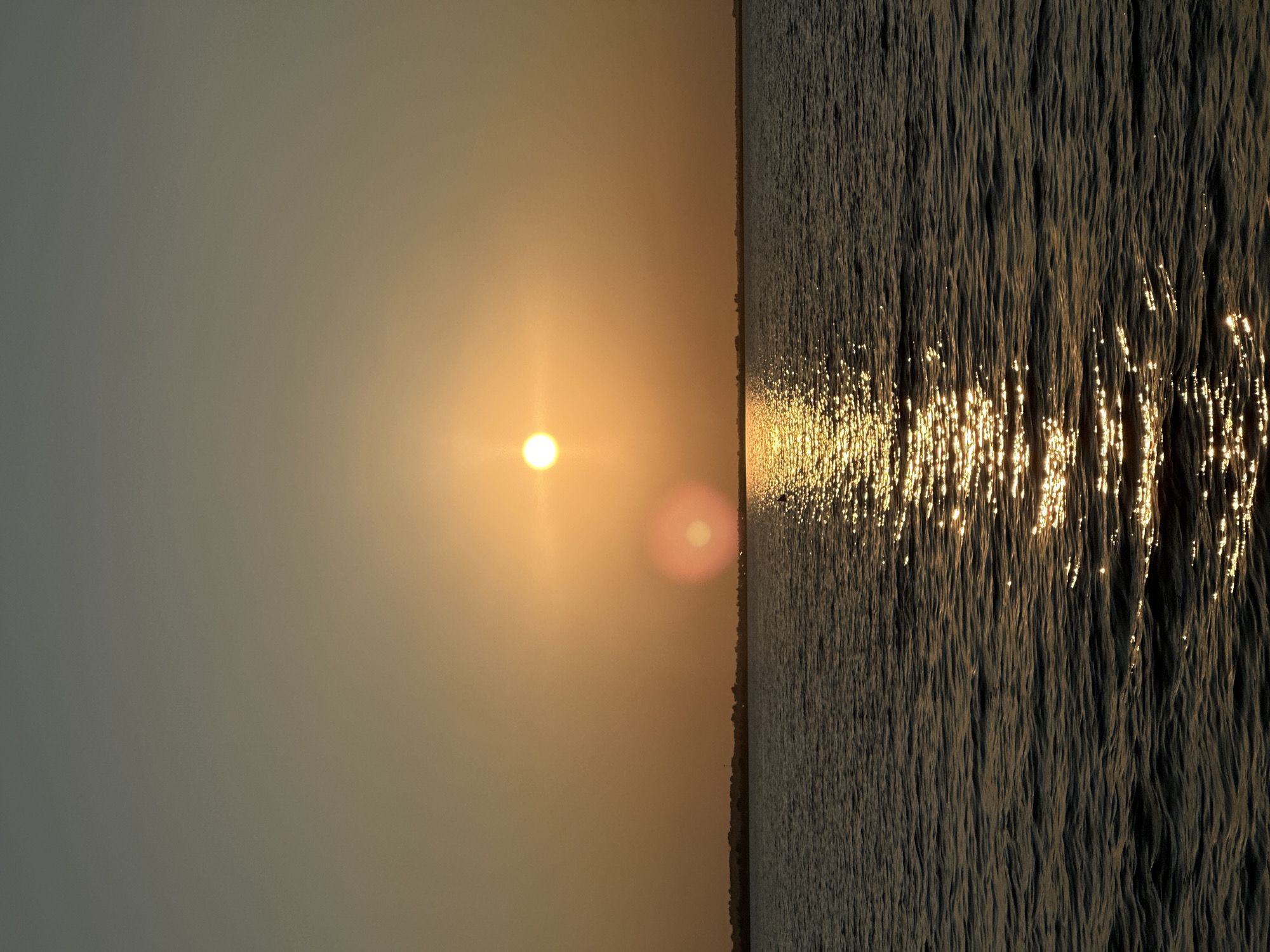 A wide shot captures a sunset over a body of water, with a silhouetted treeline visible on the horizon. The sky is a gradient of warm oranges and pale grays. The sun, a bright orb, is positioned slightly off-center, casting a shimmering reflection on the dark, rippling water. There is a faint lens flare visible near the sun. A small, dark silhouette, possibly a boat or a person, is visible in the water, positioned in the lower center of the frame. The scene is tranquil and serene, characteristic of the natural beauty found around Lake Borovoe, near Kokshetau, Kazakhstan.