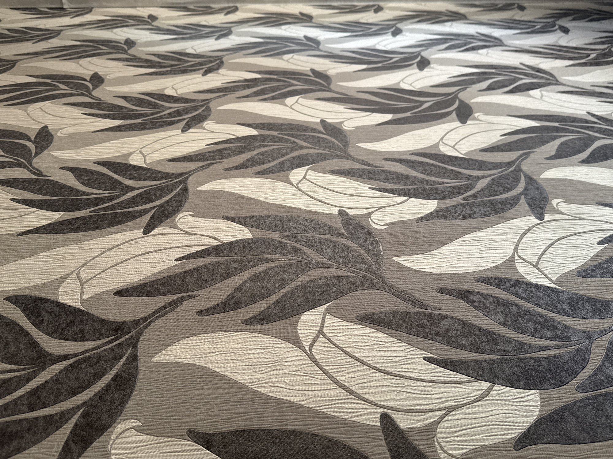 The image shows a close-up of wallpaper with a repeating pattern of stylized leaves. The leaves are rendered in dark grey and light beige, with a textured effect. The background also has a subtle texture, creating a sense of depth. The lighting suggests a slightly angled light source, casting soft shadows that enhance the dimensionality of the pattern. There are no people or any other discernible objects in the image, only the wallpaper itself. The location is identified as Bishkek City, Kyrgyzstan.