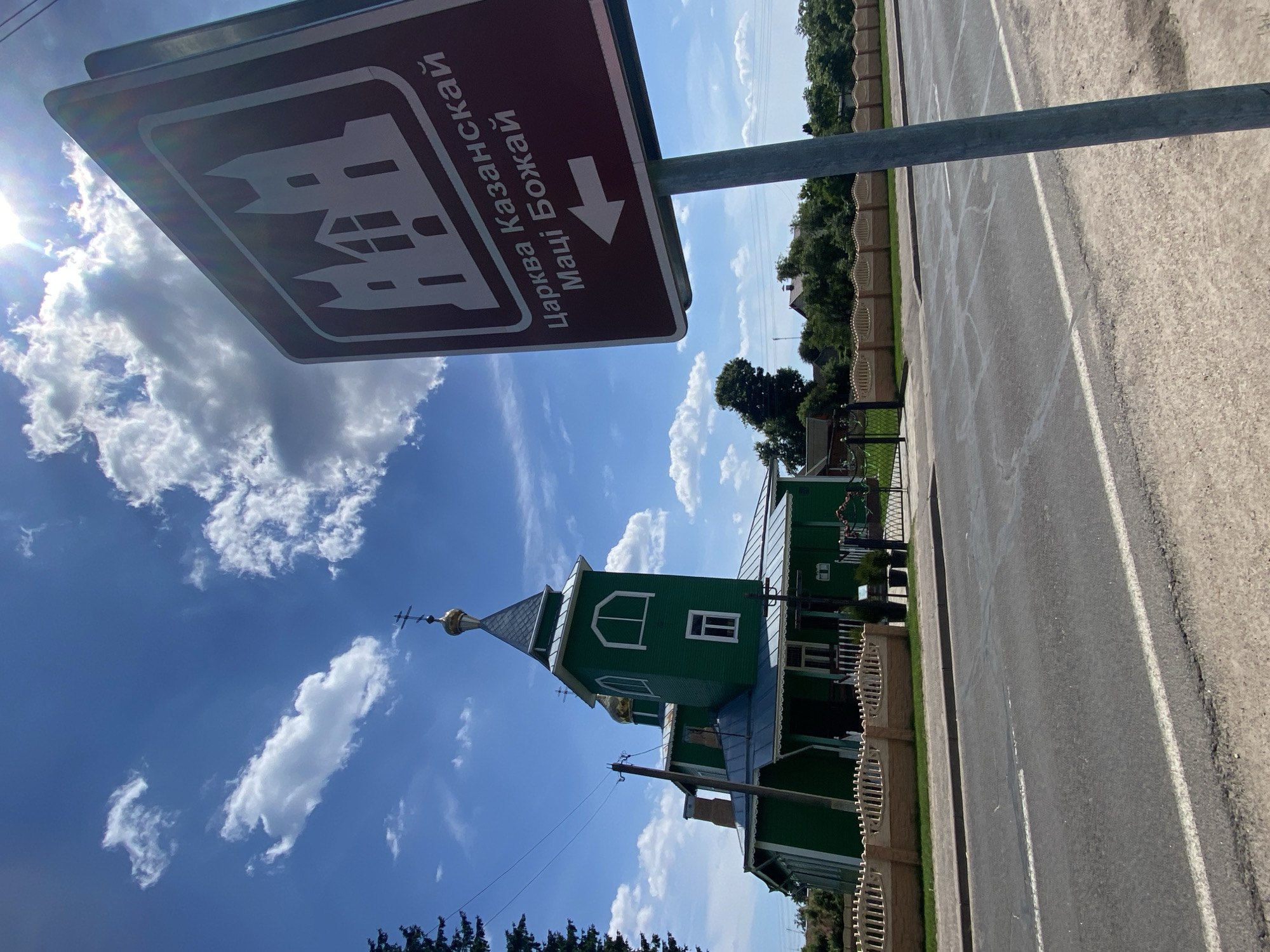 The image depicts a road sign indicating the Church of the Kazan Icon of the Mother of God in Parechcha, Belarus. The sign, a dark red rectangle with a white outline of a church and white text, is mounted on a gray metal pole. The text on the sign reads "Царква Казанскай Маці Божай" and features a white arrow pointing to the left. In the background, a green wooden church with golden domes and a tall steeple is visible. The church is surrounded by a decorative fence and greenery. The sky above is blue with scattered white clouds, and the sun is shining brightly. The road in the foreground is asphalt with white lane markings.