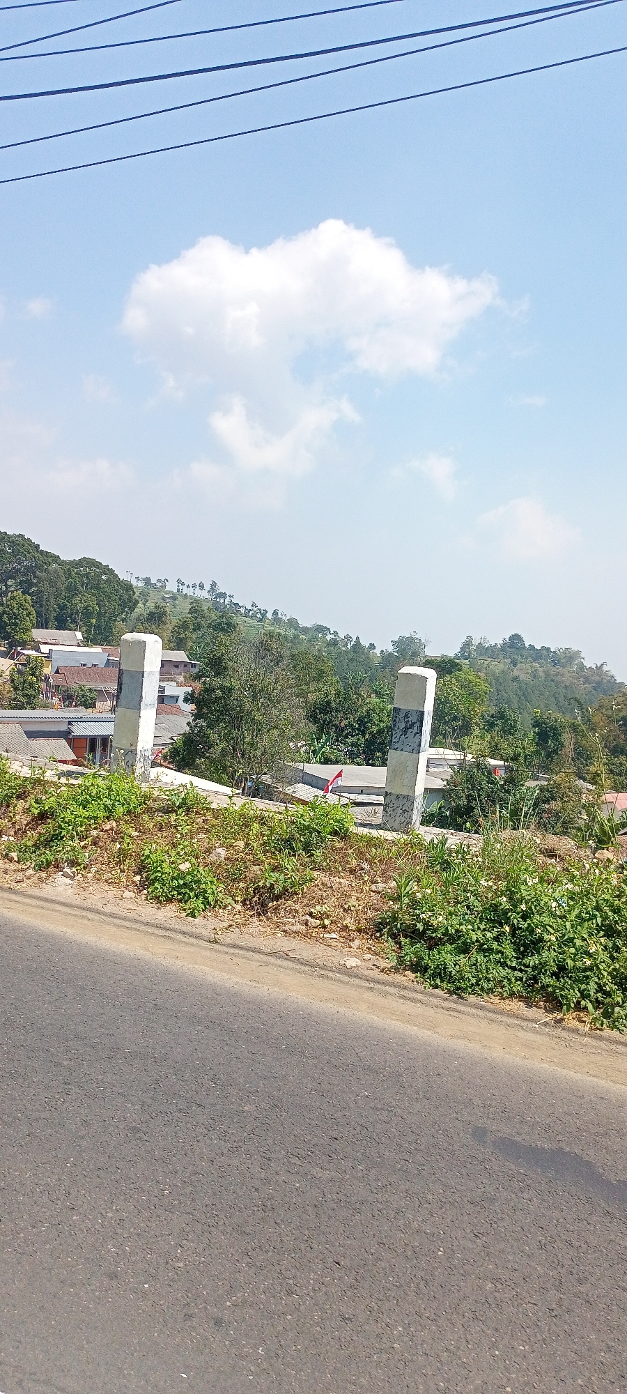 The image shows a rural landscape with a road in the foreground and a village nestled in the hills in the background. The sky is blue with scattered white clouds. There are power lines running horizontally across the top of the frame. Two concrete posts, one on each side of the road, are partially visible in the midground. These posts appear to be bollards or markers, with a distinct black and white striped pattern. The road is paved with asphalt and shows signs of wear.

The village in the background consists of several houses with red and grey roofs, surrounded by lush green trees and vegetation. The terrain is hilly, with some areas appearing to be terraced, suggesting agricultural activity. A small flag, possibly the Indonesian flag given its red and white colors, is visible on a pole in the village. The overall impression is that of a serene, rural setting in a tropical or subtropical climate. Based on the description of Cembor, Indonesia, this scene is consistent with the geography and typical appearance of villages in such regions.
