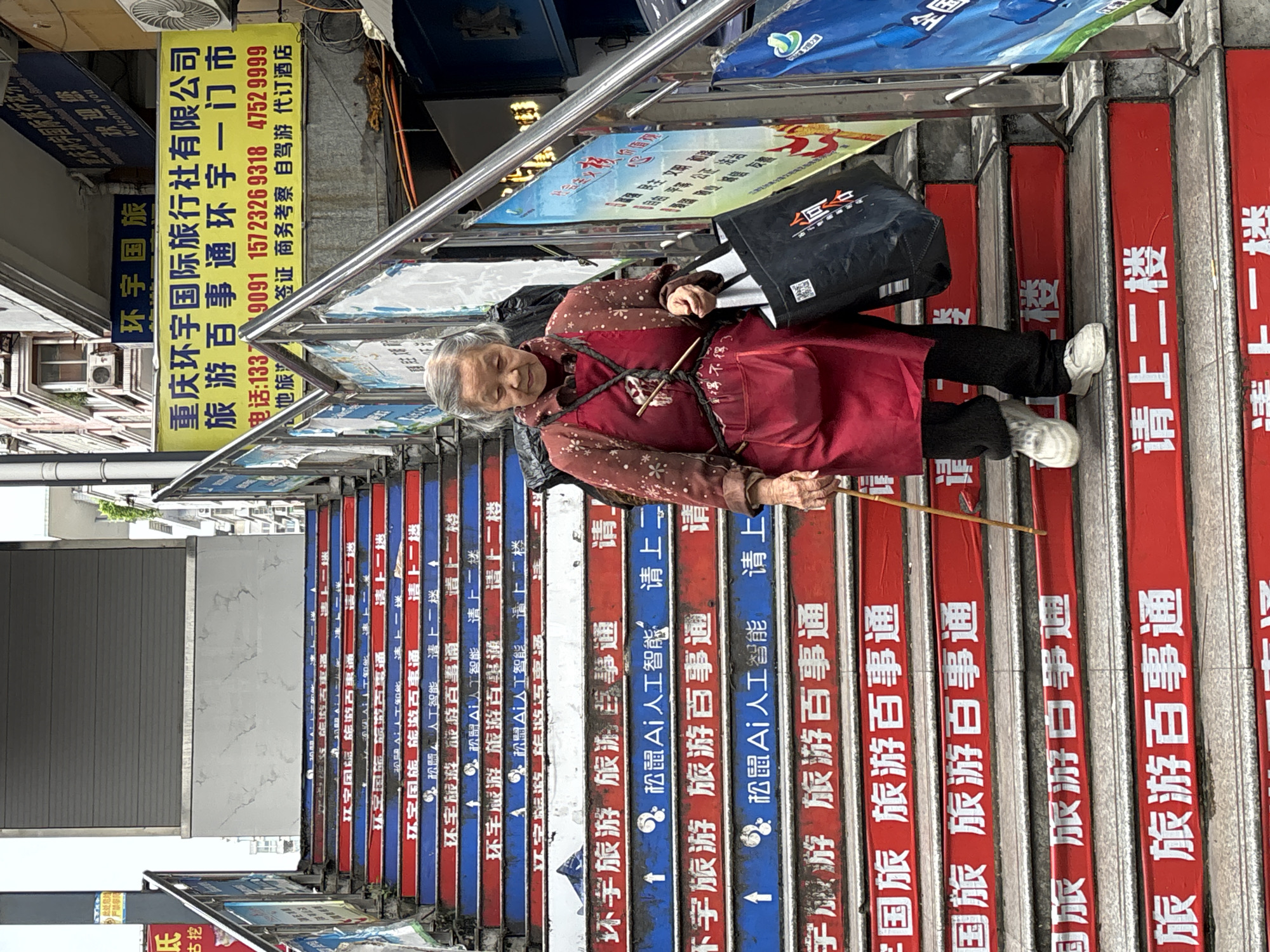 The image shows an elderly woman in Jiangjin, China, ascending a set of stairs. She is dressed in a red tunic over a patterned brown blouse, black pants, and white sneakers. She carries a black bag over her shoulder and uses a wooden stick for support. The stairs are painted with alternating red and blue stripes, each step bearing Chinese text advertising "Huan Yu Travel," accompanied by a logo and text denoting "AI intelligence". Metal handrails line the sides of the stairs, with advertisements placed alongside them. Above the stairs, a yellow sign advertises "Chongqing Huan Yu International Travel Agency Co., Ltd." The city of Jiangjin is assumed from the presence of the Chinese characters which suggest a locality.