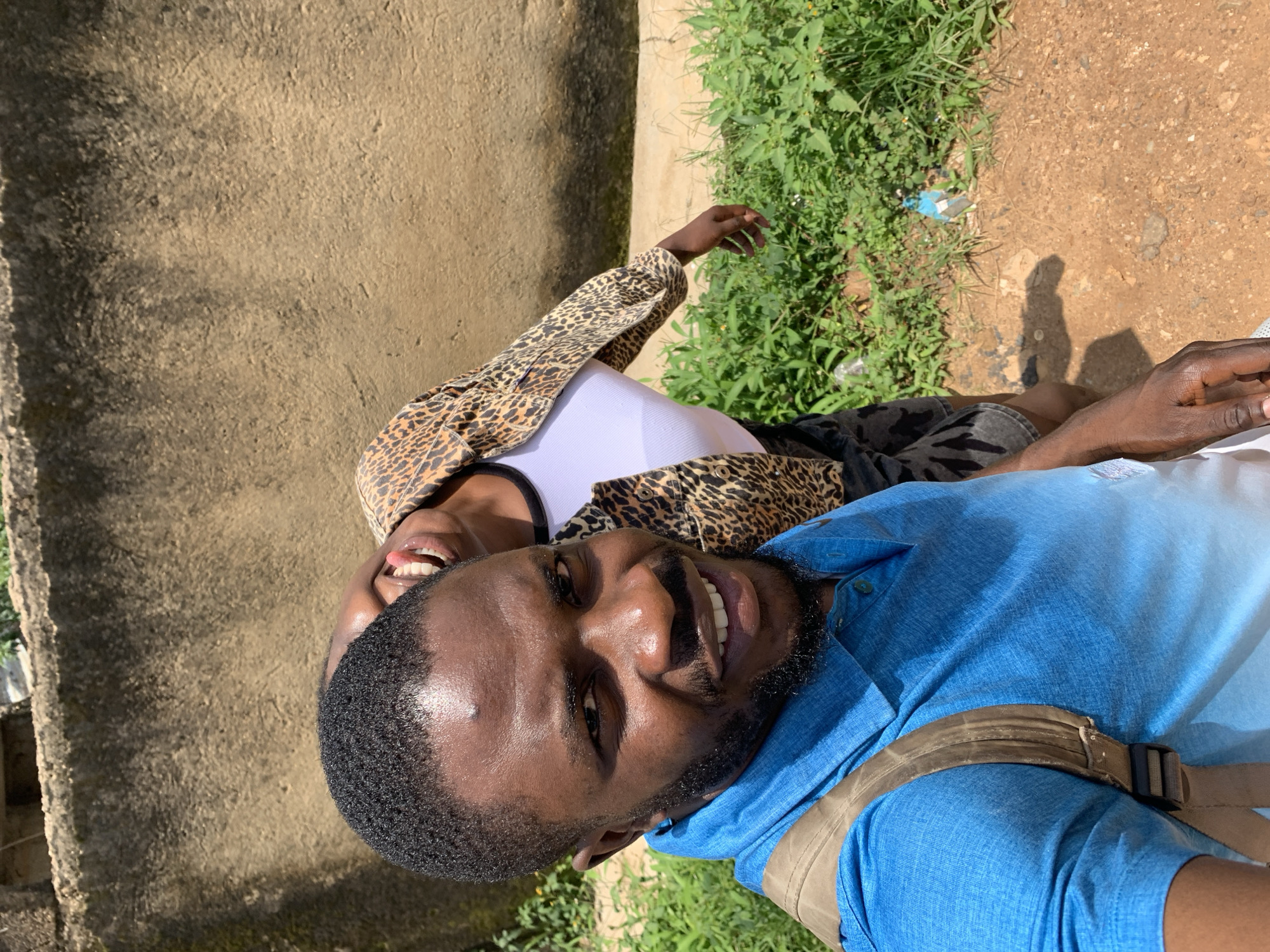 Two individuals are posing for a selfie outdoors with a textured, brown wall in the background. The person in the foreground, a man, is wearing a blue collared shirt and a beige backpack strap is visible over his shoulder. He has dark skin, a beard, and is smiling directly at the camera. Behind him, a second person, whose head is tilted back and mouth is open in a laugh, is visible. This second person is wearing a white top and a leopard-print jacket. They are standing in front of a patch of green vegetation and a dirt path. A small blue bottle is lying on the ground near the vegetation. The scene suggests an informal, outdoor setting, possibly in the city of Rafin Par, Nigeria, given the context.