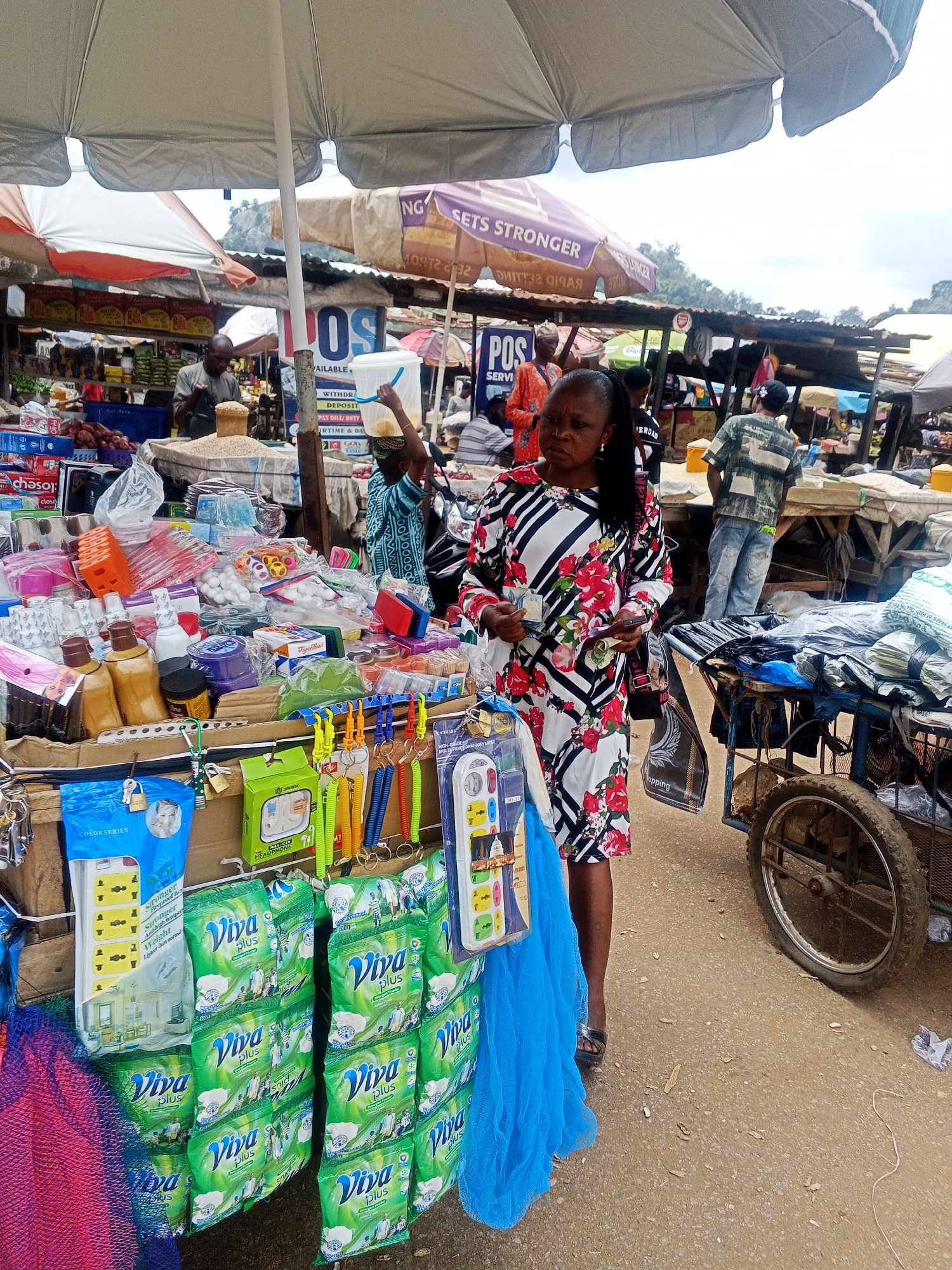 A bustling outdoor market scene is depicted in Kabba, Nigeria, under a canopy of weathered umbrellas. In the foreground, a woman in a floral-patterned dress stands amidst a vibrant display of goods. She holds cash in her left hand and a mobile phone in her right. To her left, a variety of items are neatly arranged, including stacks of green "Viva Plus" detergent packets, electrical outlets, and colorful keychain accessories. A blue mesh material is draped on the lower left. Behind her, a market stall overflows with goods, and a handcart laden with items is parked to the right. In the background, other vendors and shoppers are visible, some tending to their stalls, while others are engaged in transactions. The environment appears to be an open-air market with stalls constructed from corrugated metal and wood. The ground is a dusty, dirt surface. The overall impression is one of active commerce and daily life in a Nigerian market.