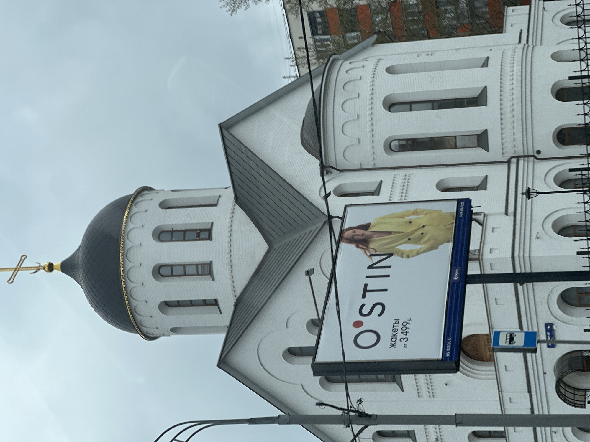 The image shows a church building with a black dome and a golden cross. A large advertising billboard is affixed to the building, advertising the clothing brand "O'STIN" and jackets for 3499 rubles. A street sign with the name "ВНИКИ 124" is visible below the billboard. The image was taken in Moscow, Russia.
