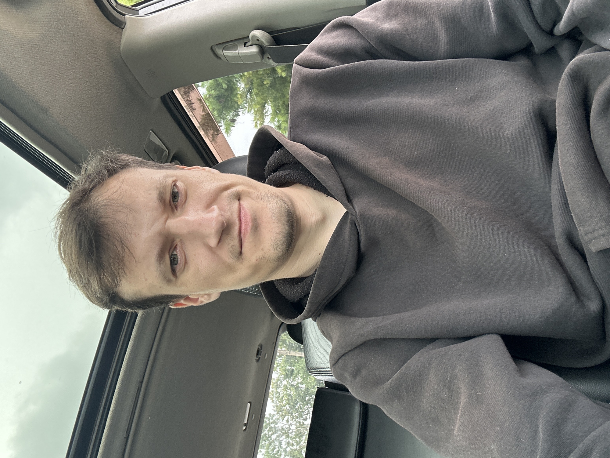 A man is sitting in the driver's seat of a car, looking at the camera and smiling slightly. He is wearing a dark gray hooded sweatshirt. The car's sunroof is open, and the sky above is overcast. Trees and foliage are visible through the car's windows. The image appears to be taken from inside the car, with the man taking a selfie. There are no visible indications of the location being in the city of Makiv, Ukraine.