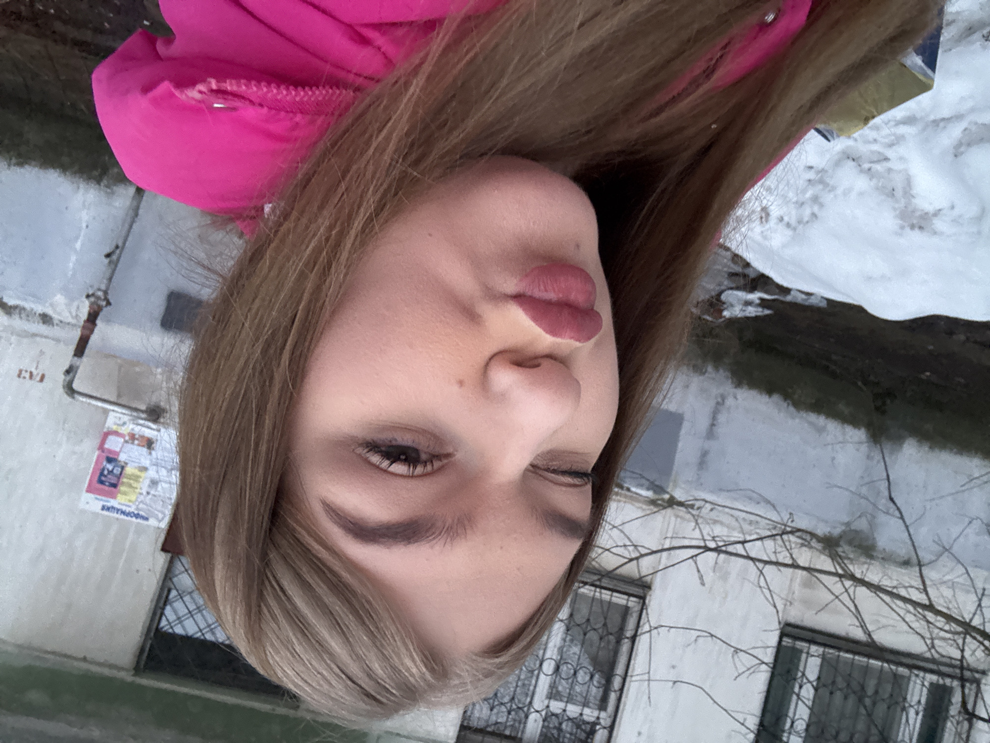A young woman is posing for a selfie outdoors. She is wearing a pink jacket and has long, light brown hair. She is puckering her lips and winking. The background shows a building with barred windows, a poster on the wall, and some snow on the ground. Bare tree branches are visible as well. The location appears to be in a residential area of the city of Chaikovsky, Russia, based on the architectural style of the building and the presence of the Russian-language poster.