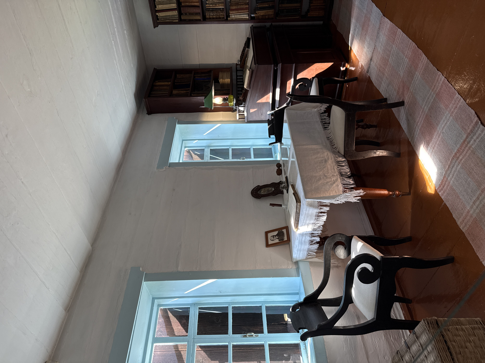 The image depicts an interior scene of a room, likely a historical dwelling or museum exhibit, in the city of Shushenskoye, Russia. The room features a wooden floor and white-painted log walls, with exposed horizontal beams forming the ceiling. Two light blue framed windows with wooden panes allow sunlight to stream into the room, casting long shadows.
In the center of the room, a wooden table is draped with a white tablecloth that has a fringed edge. On the table are several objects, including an open book, a framed photograph of a man, an inkwell with a quill pen, and a small clock. Two chairs are positioned at the table: a dark, ornate wooden chair with a light-colored upholstered seat is in the foreground to the left, and another similar chair is to the right.
To the right of the table, a dark wooden lectern-style desk is visible, topped with books and papers. A green-shaded oil lamp sits on the desk, its light illuminating the area. Behind the desk are two tall, dark wooden bookshelves filled with numerous books. Another similar bookshelf is partially visible on the far right side of the image. A long, woven rug with a checkered pattern of pink, white, and grey runs diagonally across the floor in front of the desk. A woven basket is situated in the lower left corner of the frame. The overall impression is that of a study or personal workspace, preserved to reflect a past era.
