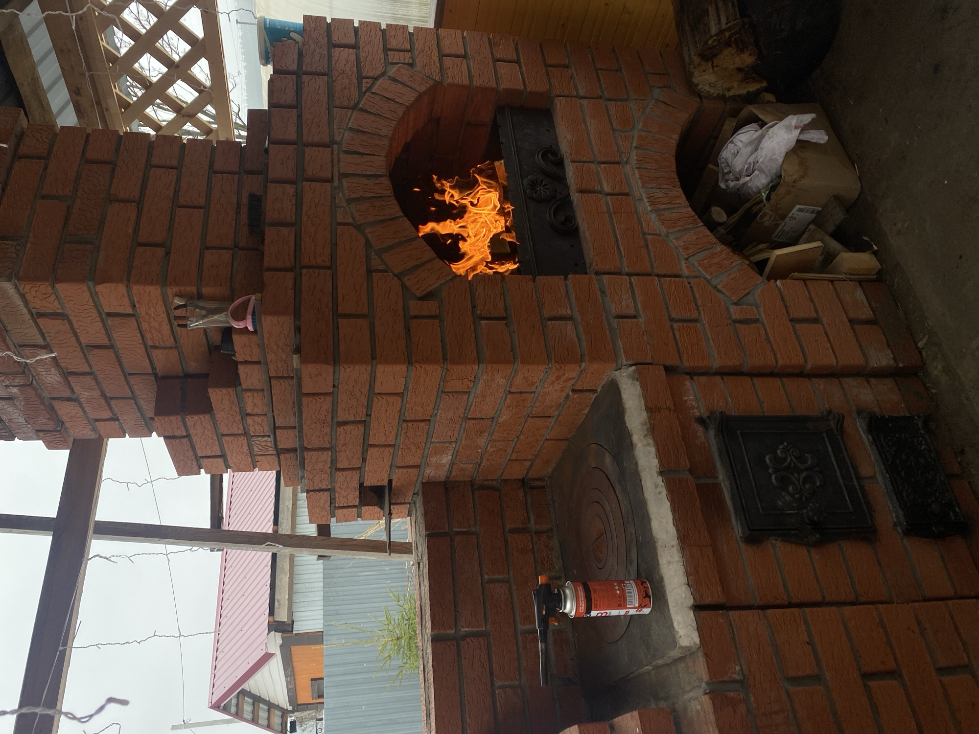 The image shows a brick oven with a fire burning inside. The oven is located outdoors, possibly in a backyard. There is a metal door on the front of the oven. To the right of the oven opening there is an opening for storing wood and kindling. Under the opening for the fire, there is a burner resting on a slab of stone. In the background, a wooden structure, a fence, and the roof of a building is visible. The location is Chaikovsky, Russia.