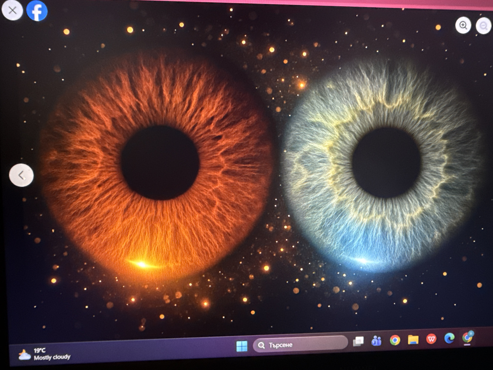 The image is a photograph of a computer screen displaying a desktop wallpaper. The wallpaper features two large images of human irises, one orange-brown and one blue-grey, set against a dark background with numerous small, blurred light points, reminiscent of stars or bokeh. A bright, lens-flare effect is visible at the bottom of each iris.
Icons and a taskbar are visible at the bottom of the screen. The taskbar shows weather information ("19°C Mostly cloudy"), a search bar, and various application icons. In the top left corner, there are two application icons, including a Facebook logo. In the top right corner, there are two magnifying glass icons.