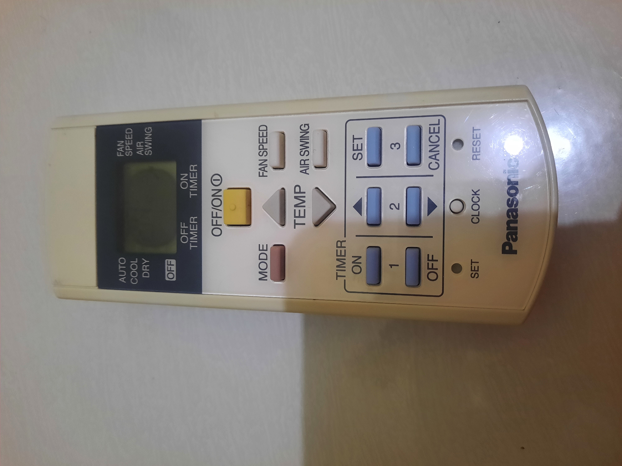 A close-up, eye-level shot shows a beige Panasonic air conditioner remote control lying on a light-colored, textured surface, possibly a table or floor. The remote is positioned slightly off-center, with its top edge angled towards the right. The remote is rectangular with rounded corners and a digital display screen at the top, which is dark. Below the screen are several buttons. On the left side, vertical text reads "AUTO," "COOL," and "DRY," with a horizontal "OFF" button beneath "DRY." To the right of the digital display, vertical text reads "FAN SPEED," "AIR," and "SWING." Below these, horizontal text reads "OFF" and "ON TIMER." A prominent "OFF/ON" button with a small circular symbol is located below the display. Further down, "MODE" is to the left, with two arrow buttons ("TEMP") above and below it. To the right of "TEMP" are "FAN SPEED" and "AIR SWING" buttons. The bottom portion of the remote features "TIMER ON" with buttons labeled "1" and "2" arranged vertically on the left, and "SET" with a button labeled "3" on the right. Below these are "OFF" and "CANCEL" buttons. At the very bottom, "SET," "CLOCK," and "RESET" are labeled with circular indicators above them. The "Panasonic" logo is printed in black on the lower right side of the remote. The lighting creates a slight glare on the bottom right of the remote and the surface it rests on. The image context does not provide any information about the city of Lhokseumawe, Indonesia.