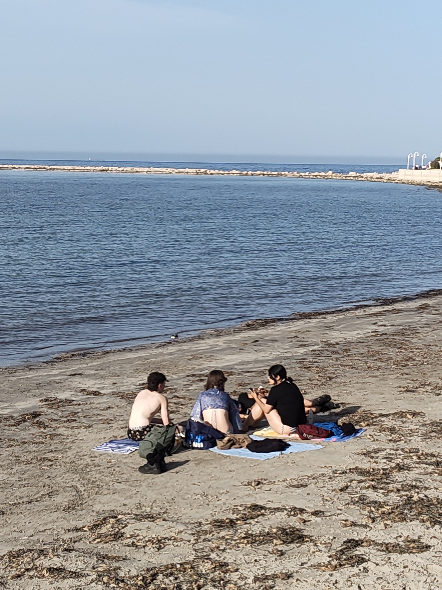 Three people are sitting on a light blue towel on the beach, facing the sea. The person on the left is a male with bare torso and dark colored bottoms. He is sitting, partially turned towards the group. The person in the middle has on a cropped blue shirt with what appears to be a bikini or swimsuit bottom. The person on the right is wearing a dark colored t-shirt. They appear to be eating and holding a mobile phone. Clothing and backpacks are scattered around them on the towel. The beach is covered in seaweed. In the background, the sea is calm and blue. A breakwater stretches across the horizon line. On the right side of the image, is a paved area with light colored concrete, possibly a sidewalk or path. The location is likely in Dénia, Spain.