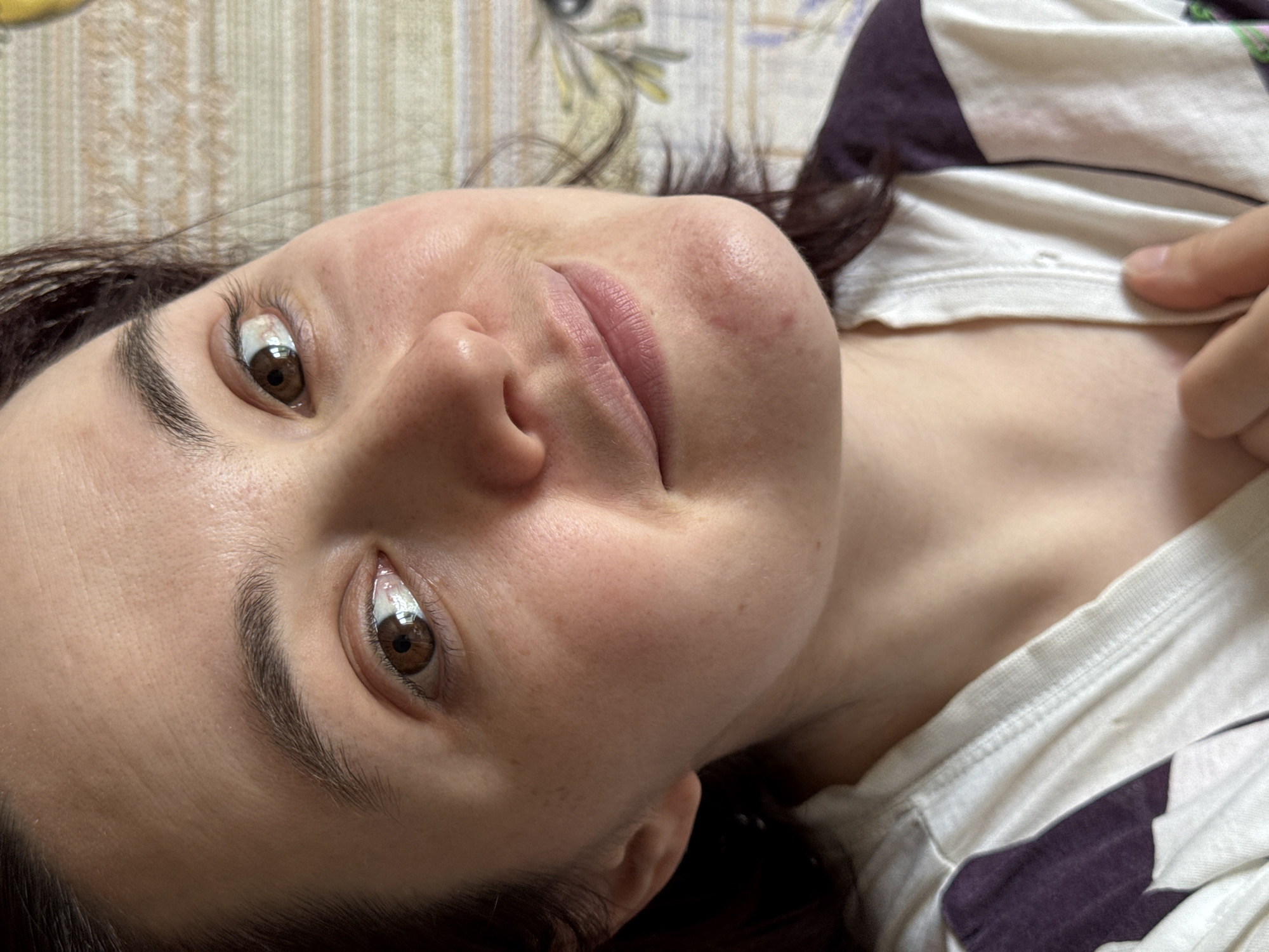A woman is lying on a bed. She has brown eyes and dark hair. She is wearing a white and purple shirt. The background is a light-colored patterned fabric. The location is not identifiable as Izhevsk, Russia, based on the image.