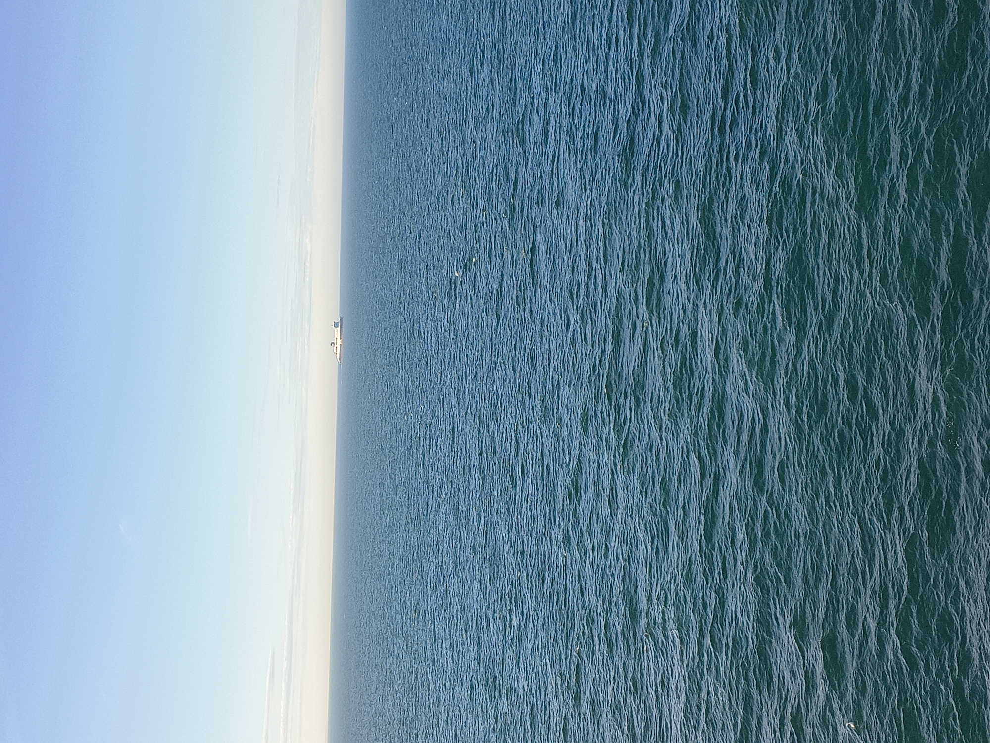 An image taken from a high vantage point shows a wide expanse of the sea under a clear blue sky with a thin band of clouds on the horizon. The water is a deep blue-green, with small ripples and waves visible across the surface. In the distance, a large white ship is visible on the horizon, appearing as a small speck against the vastness of the sea and sky. There are no people or any other discernible objects in the scene, creating a sense of isolation and open space. The location is identified as Unknown, United Kingdom.