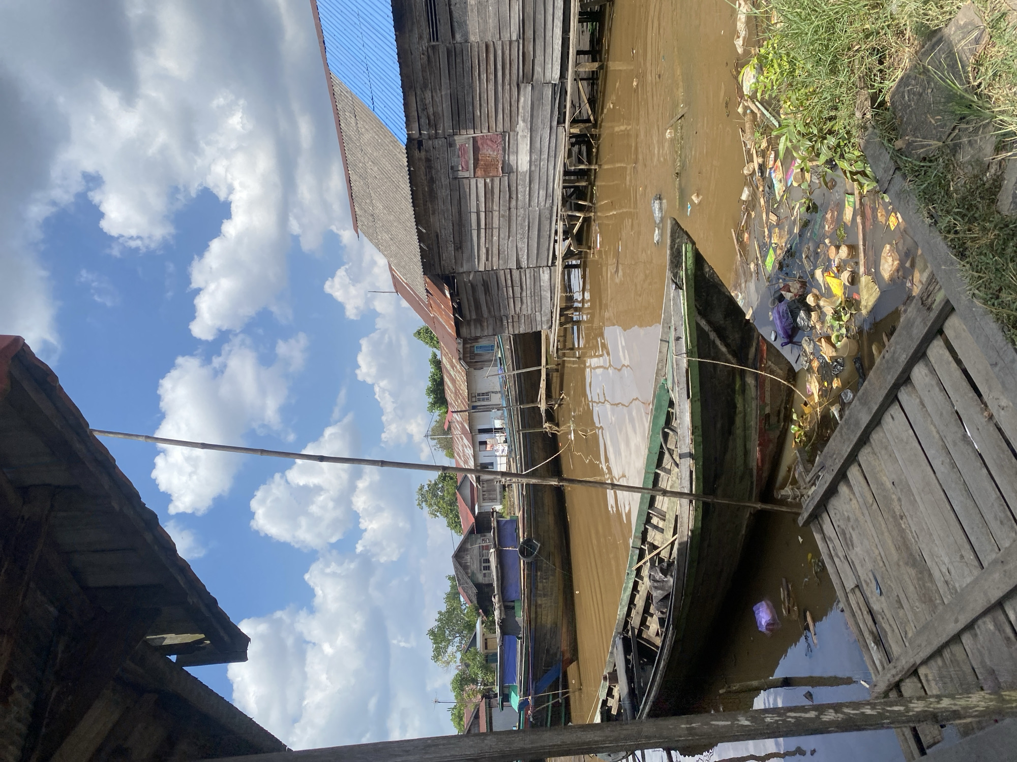 In the city of Berangas Timur, Indonesia, the image captures a view of a brown, muddy river with several wooden boats docked along its bank. The boats vary in size and condition, with one in the foreground appearing weathered and carrying a dark cover. The river is heavily polluted with various forms of trash, including plastic bags, bottles, and other debris, floating on the surface and accumulated near the bank.
Along the riverbank, wooden stilt houses are visible, some with corrugated metal roofs, one of which is painted blue. The architecture suggests a vernacular style typical of riverine communities. The overall environment is a mix of human habitation and natural elements, with lush green vegetation encroaching on the edges of the river and some trees in the background. The sky is partly cloudy, with white cumulus clouds against a bright blue expanse. A long bamboo pole is positioned vertically in the water, possibly used for mooring or navigation. The scene conveys a sense of a bustling, yet potentially struggling, riverside community.