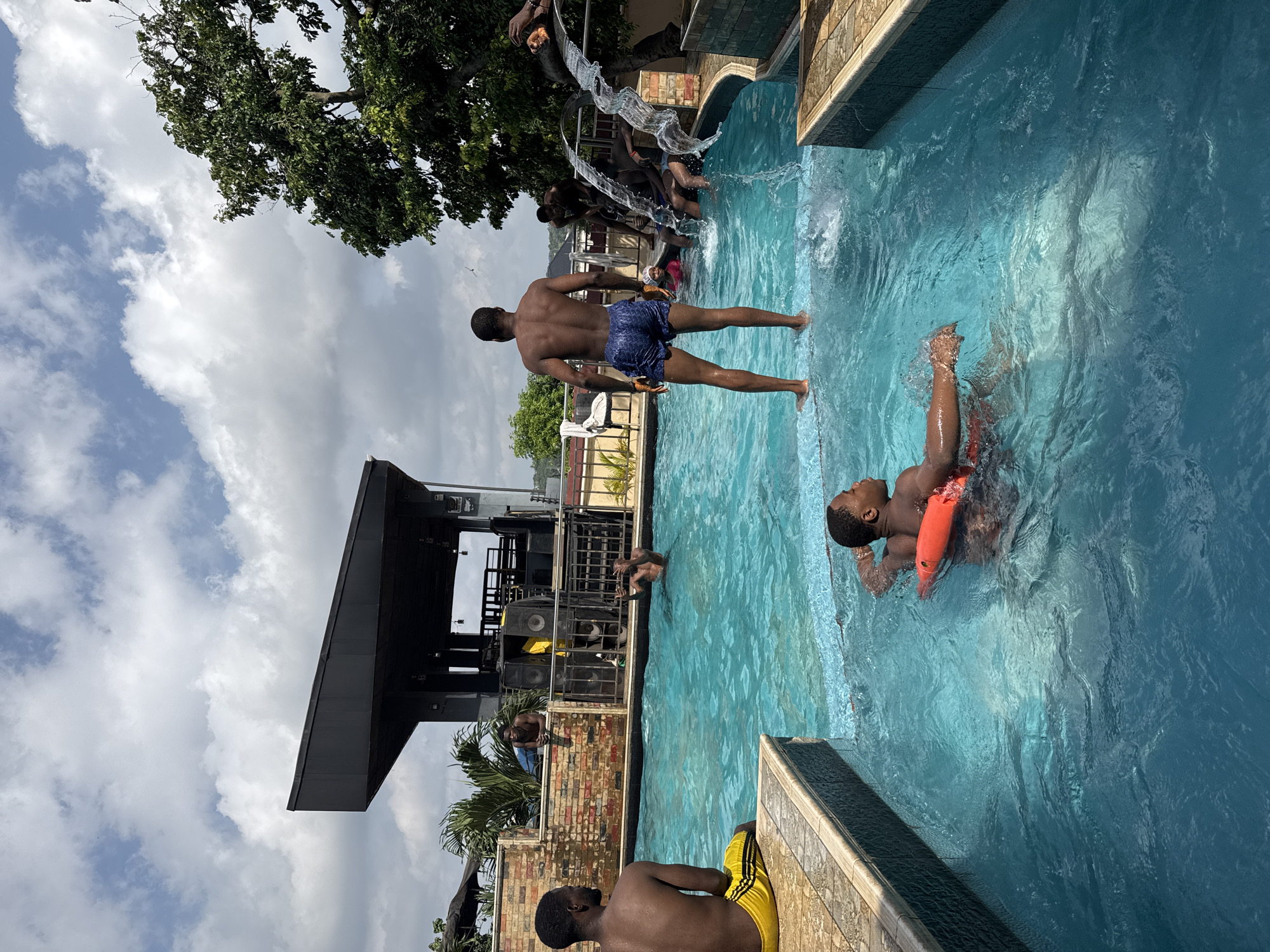 In Nkpor, Nigeria, on a sunny day, a group of young men are gathered at an outdoor swimming pool, possibly at a club or resort, situated near a building with a distinctive black, angular roof and speakers set up on a raised platform. The pool's vibrant turquoise water is surrounded by a beige tiled deck and brick walls. In the foreground, a young man wearing yellow and black swim shorts sits on the edge of the pool, facing away from the camera. In the center, another young man in blue swim shorts stands on the pool's edge, facing the water. A third young man, wearing an orange flotation device, floats in the pool closer to the foreground. Several other individuals are visible in the water and on the pool deck. One young man is partially submerged in the water, looking over his shoulder. Another man is seen with his hands raised. Water cascades from a decorative feature into the pool from the right side of the image. Lush green trees with dense foliage frame the right side of the scene, and a partly cloudy sky with white and gray clouds completes the backdrop.
