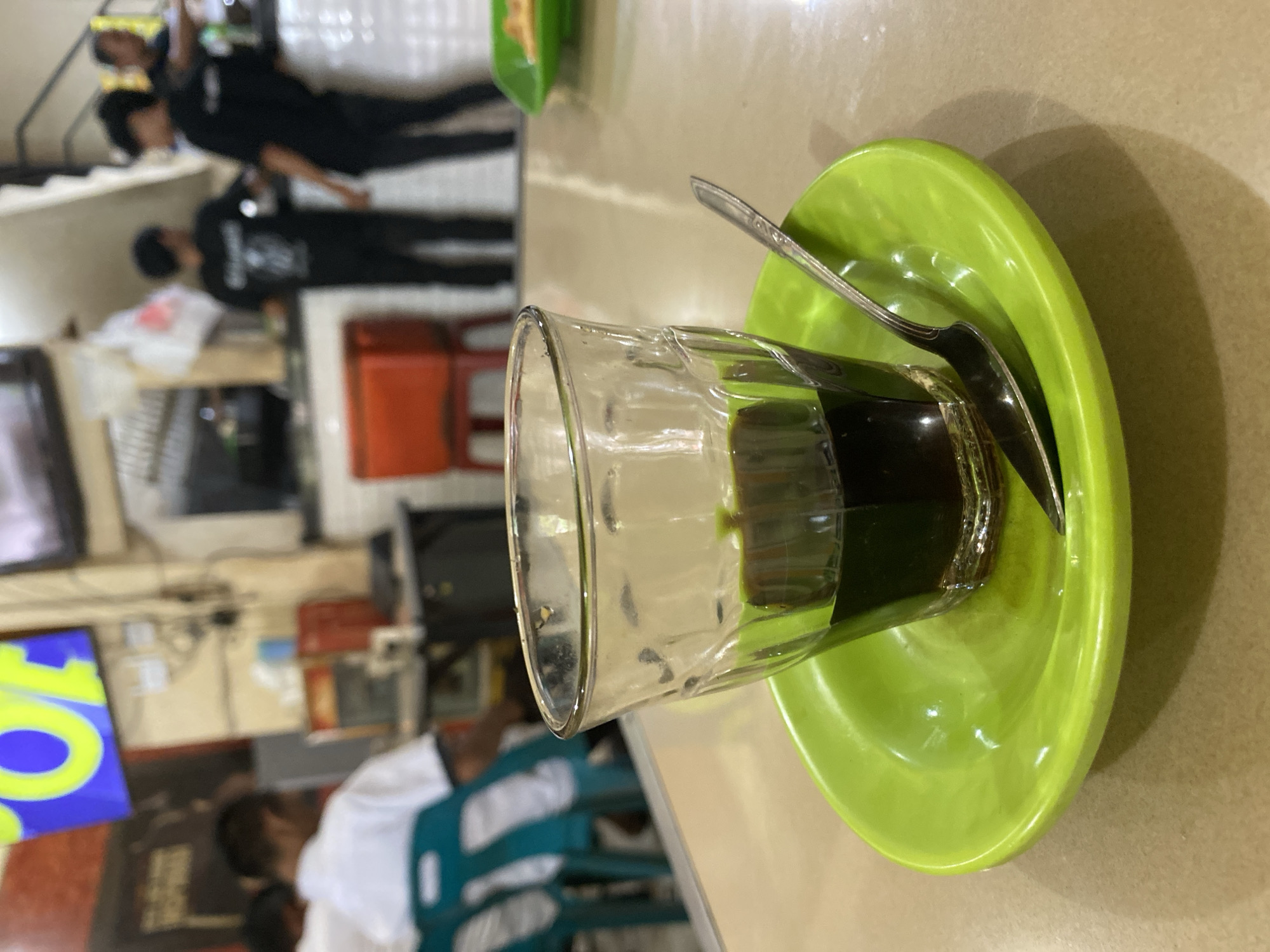 A glass of dark liquid, possibly coffee, sits on a bright green saucer with a spoon. The glass is partially filled, showing a deep brown color. The setting appears to be indoors, possibly a cafe or restaurant, with a light-colored table. In the background, several people are visible. Some are wearing black shirts and appear to be standing or moving around. One person in the foreground is wearing a white shirt. The overall lighting suggests daytime. The location is Lhokseumawe, Indonesia. There is no visible text in the image.
