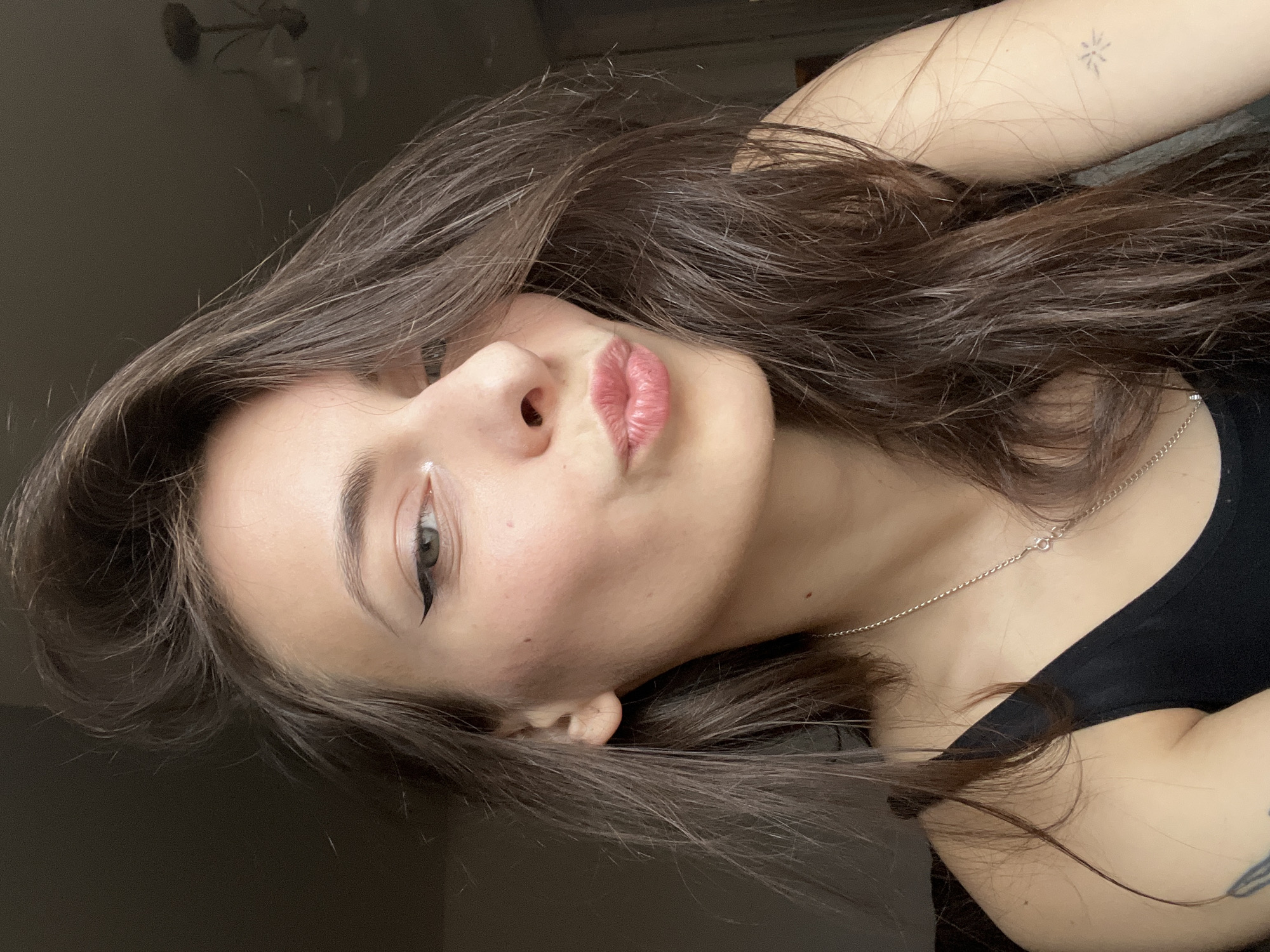 A young woman with long, brown hair is photographed from a close, angled perspective. She has fair skin, defined eyebrows, and wears eyeliner. A small, white cosmetic dot is visible on her face. Her lips are slightly pursed. She wears a thin silver necklace and a black top. A small tattoo is visible on her upper arm. The background is blurred and dark, possibly indoors. The location is not possible to determine from this image.
