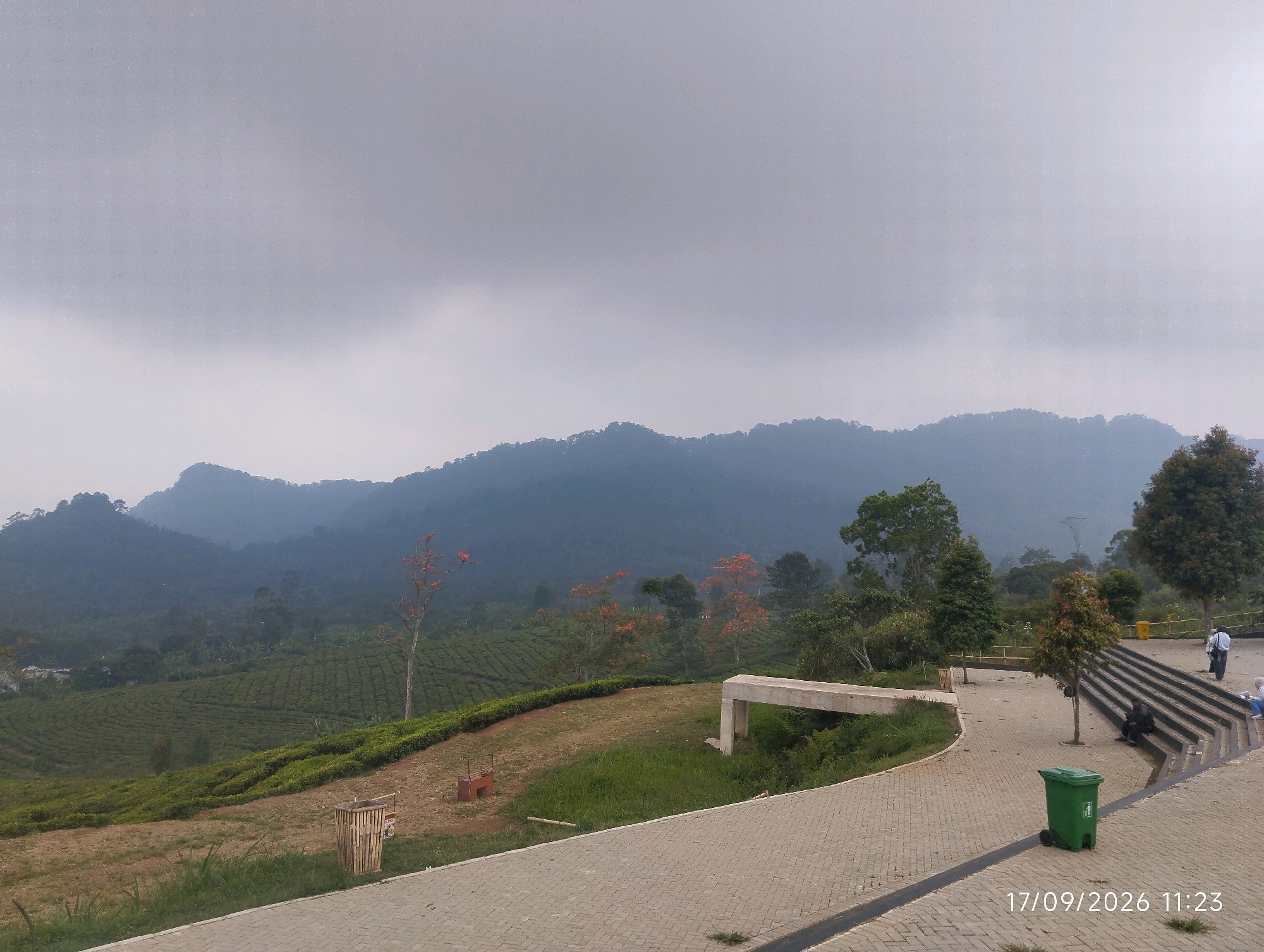 This is an outdoor, wide-angle, eye-level shot of a scenic overlook in Ciater, Indonesia, on a cloudy day. In the foreground, a paved walkway made of interlocking bricks curves from the lower right to the lower left of the frame. To the right of the walkway, a set of stone stairs leads upwards. A green trash can with wheels is positioned at the bottom of the stairs. On the stairs, two people are visible: one person in a white shirt and dark pants is standing, while another person in dark clothing is seated. Further up the stairs, other individuals are out of focus. To the left of the walkway, a small, empty wooden trash bin and a red brick structure are visible on a grassy slope. Beyond the grassy area, a vast expanse of green tea plantation stretches across rolling hills, leading to a backdrop of misty, blue-hued mountains. Several trees, some with vibrant orange flowers, are scattered throughout the landscape. A concrete structure, possibly a bench or a viewing platform, is situated on the grassy area. The sky is overcast with a muted grey hue. The timestamp "17/09/2026 11:23" is visible in the lower right corner of the image.