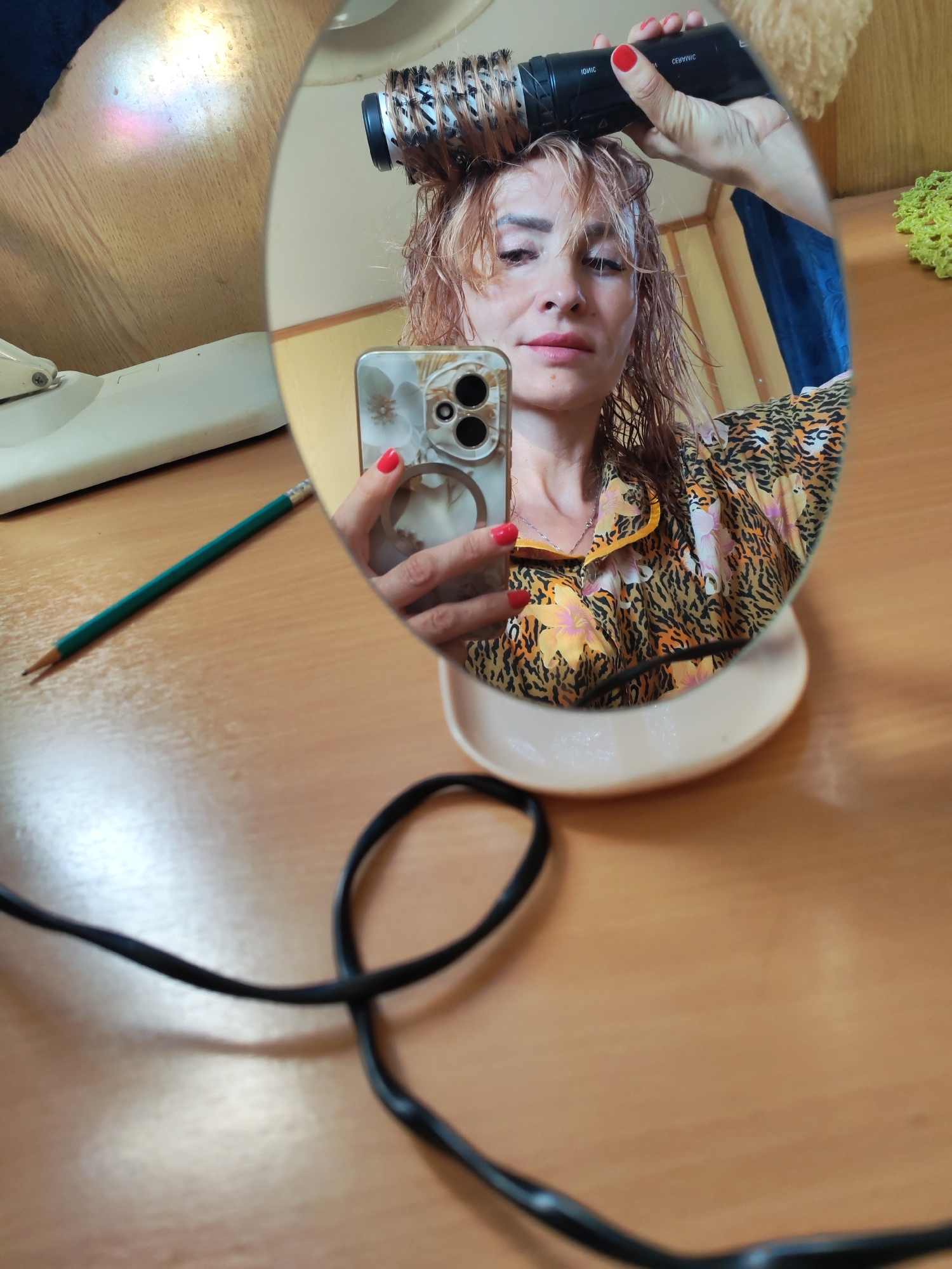 A woman is reflected in a mirror, drying her hair with a round brush and a hairdryer. She has wet, reddish-blonde hair and is wearing a leopard print and floral pajama top. Her nails are painted red. She is holding a smartphone in her left hand, which is also reflected in the mirror. A black electrical cord lies on a wooden surface in the foreground. A pencil and a white object, possibly a sewing machine base, are visible to the left. The background includes what appears to be a blue curtain and a yellow fuzzy object. The scene is set indoors, on a wooden table. The location is not specified as Krasnoe, Russia in the image.