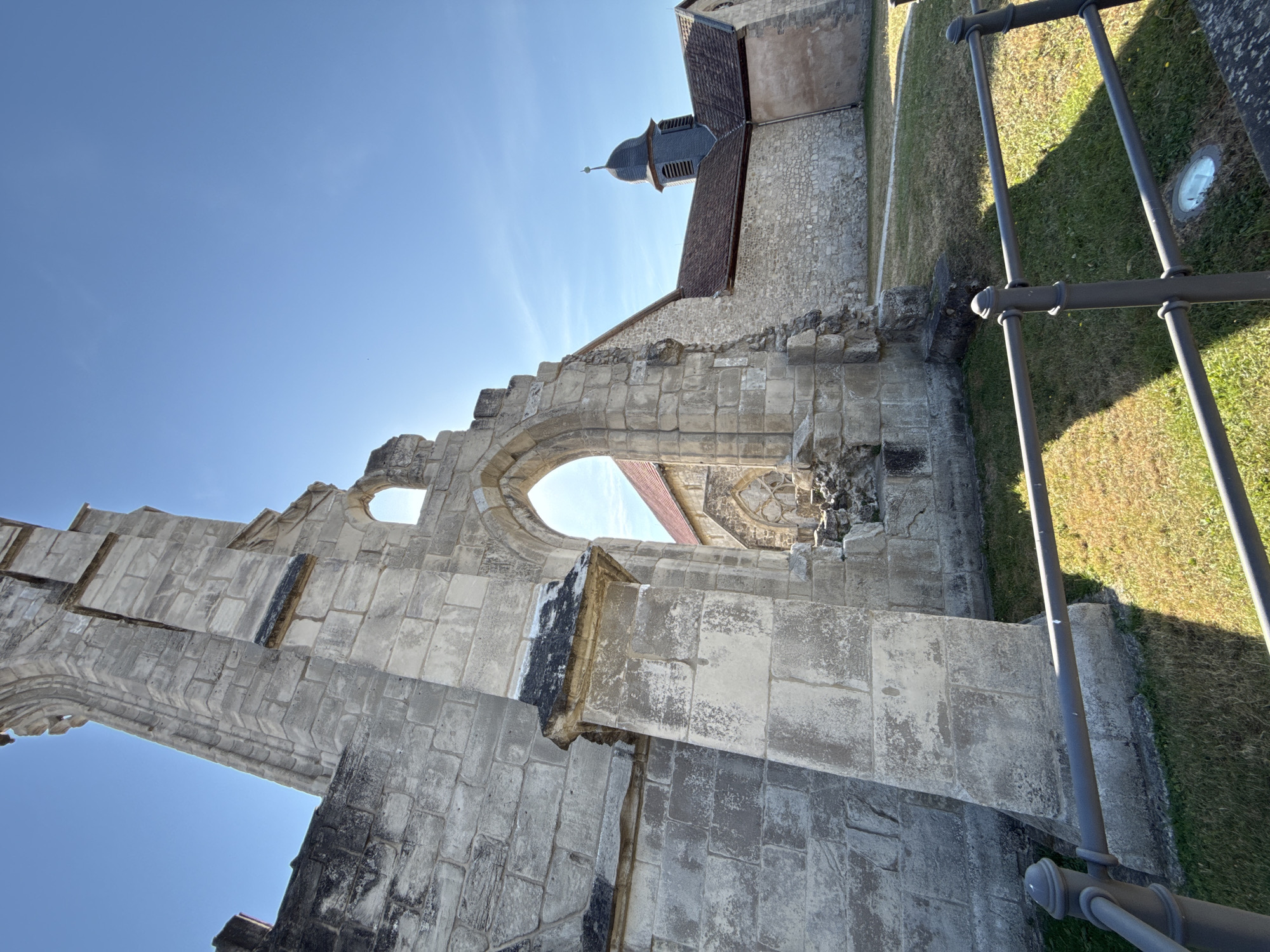 The image depicts the ruins of a stone structure, likely a historical church or abbey, situated in the town of Walkenried, Germany. The ruins are characterized by large, weathered stone blocks forming walls and an arched opening. Sunlight streams through the archway and illuminates the scene. In the background, a complete building with a tiled roof and a dark steeple is visible, suggesting it is a functioning church or an adjacent historical structure. The foreground shows a well-maintained lawn area with a dark metal fence running across it. The sky is clear and blue with a few wispy clouds. No people are present in the image.