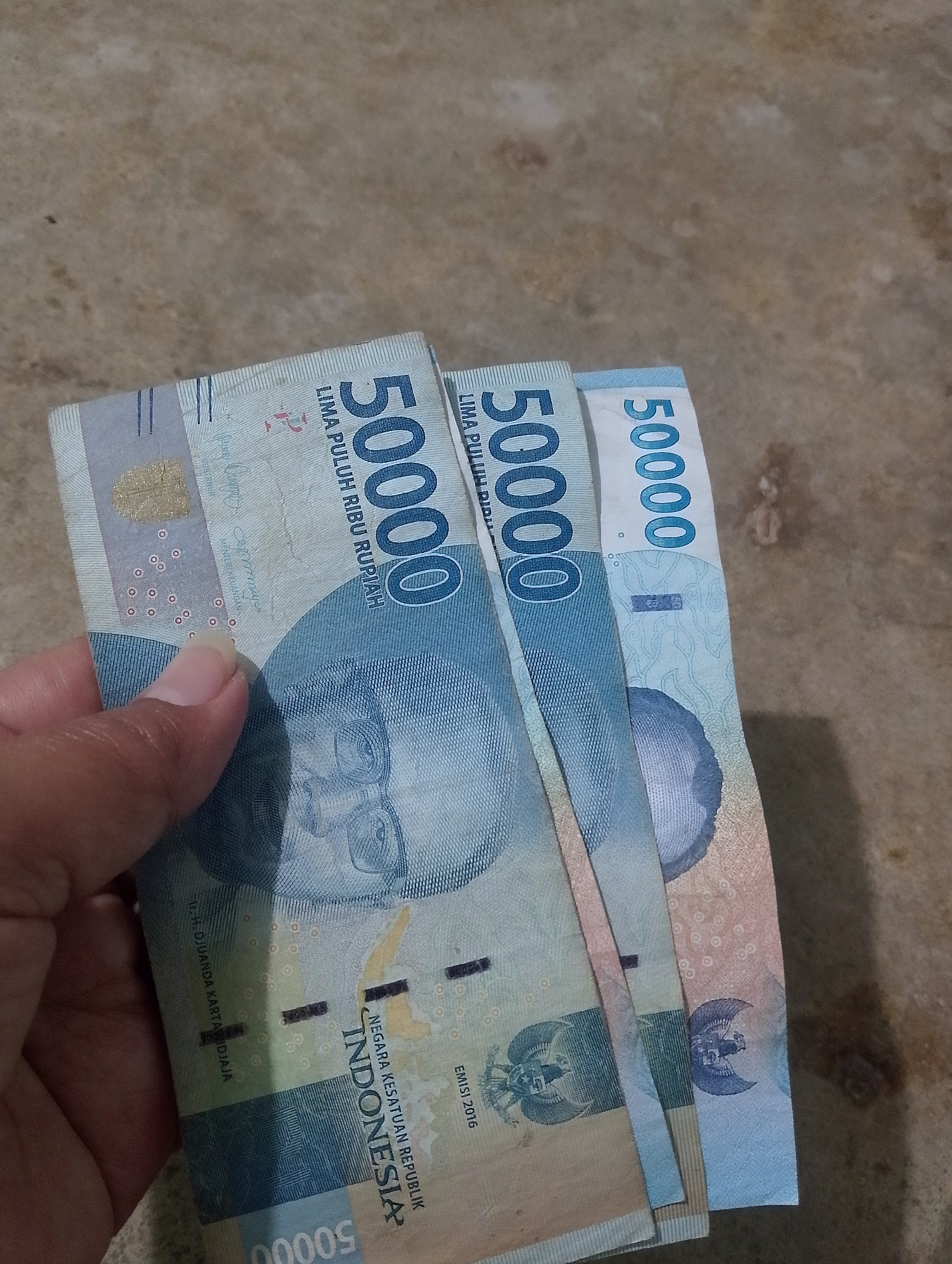 In the city of Serbelawan, Indonesia, a person's hand holds three Indonesian 50,000 rupiah banknotes. The banknotes are fanned out and partially overlap. The obverse side of the banknotes, featuring a portrait of a man and the number "50000" prominently displayed, is visible. The background consists of a textured, brownish-gray surface, possibly a floor or table. The lighting is moderate, casting some shadows. The image is taken from a slightly elevated perspective.