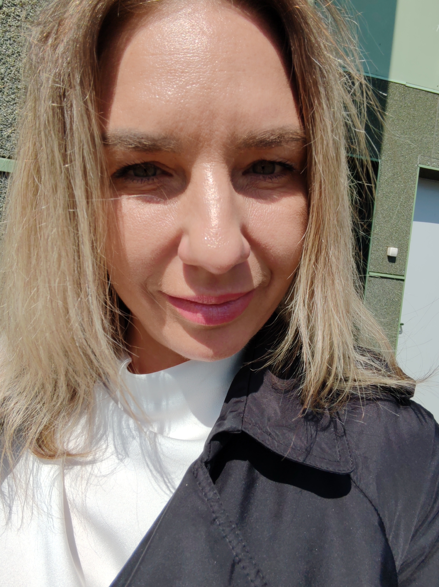 An outdoor, close-up, selfie-style photograph captures a woman with blonde hair and fair skin. She is looking directly at the camera with a slight smile. Her eyes are visible, and she is wearing a white top under a black jacket. The background appears to be a building with a green, textured facade and a window. The lighting suggests it is daytime, with sunlight casting shadows on the subject and background.