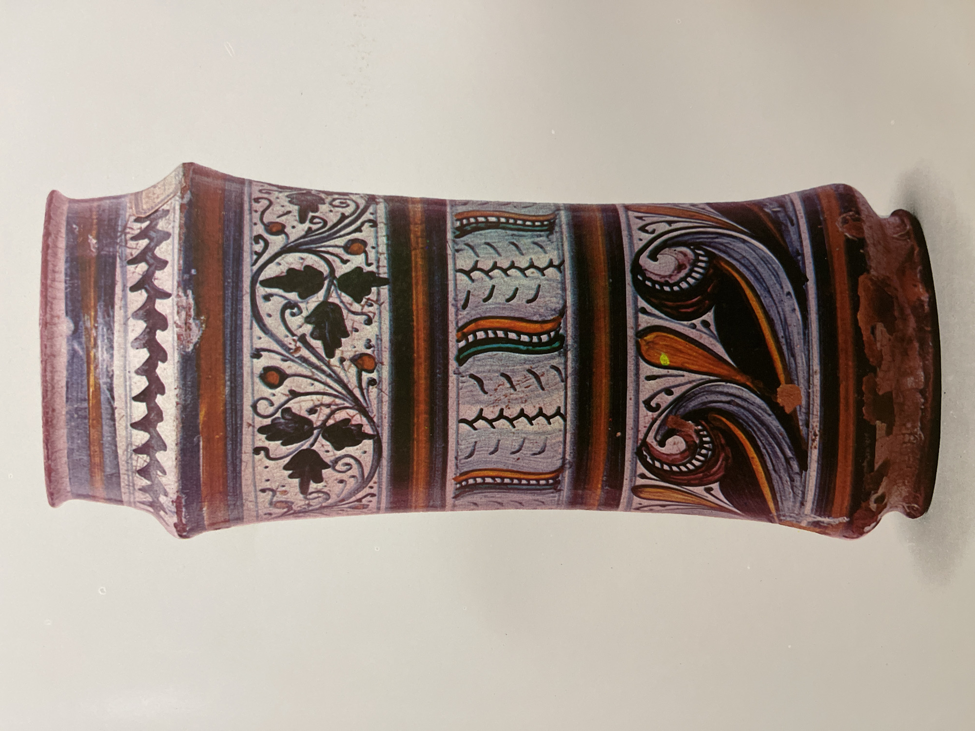 An antique or reproduction ceramic vase or cylindrical container is the focal point of the image. It rests horizontally against a plain, off-white background. The vase exhibits a series of decorative patterns, separated by vertical bands of brown and muted red hues. The decorative patterns include:
1. A row of repeating zigzag shapes.
2. A floral or botanical motif featuring dark leaves and small red berries.
3. A pattern of abstract lines with accents of orange, green, and turquoise.
4. A swirling, stylized pattern with orange, black, white, and blue elements.
The ends of the container appear to have flared lips or rims, with one end displaying some damage or wear. The container's surface also shows some minor imperfections or signs of age.
No specific location in Almaty, Kazakhstan, or any people, actions, or interactions are depicted in the image.