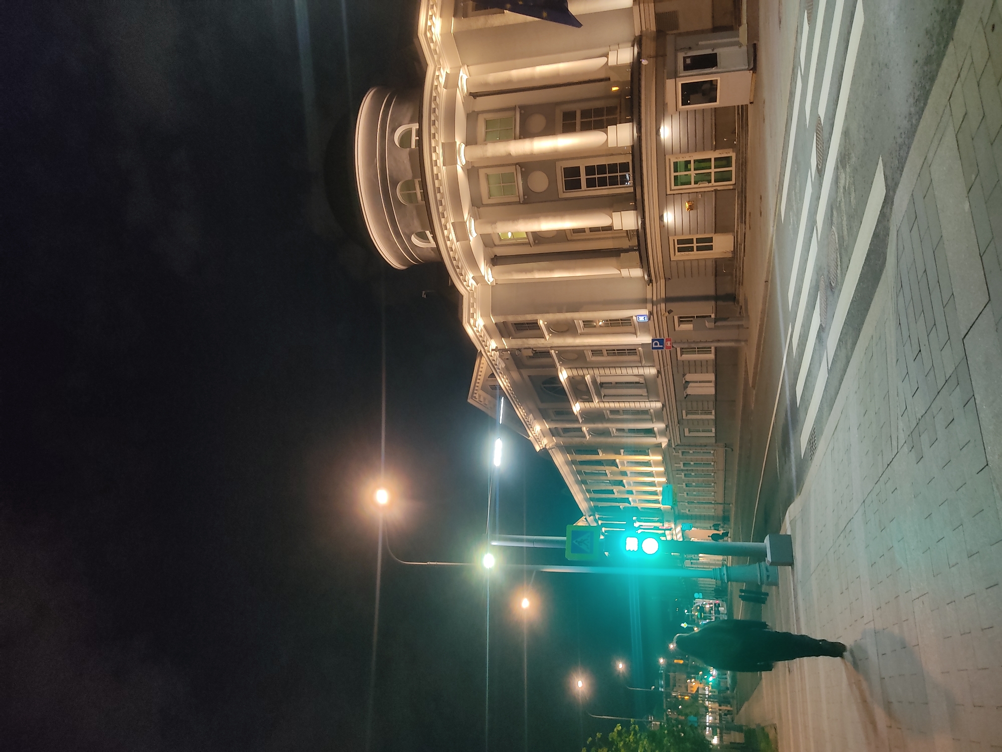 The image depicts a nighttime scene in Moscow, Russia. A pedestrian is walking across a crosswalk with zebra stripes, illuminated by streetlights and a green traffic signal. A large, ornate building with columns and illuminated windows stands adjacent to the crosswalk. The background is dark, suggesting it is late at night. The environment is urban, with a mix of architectural and infrastructural elements.