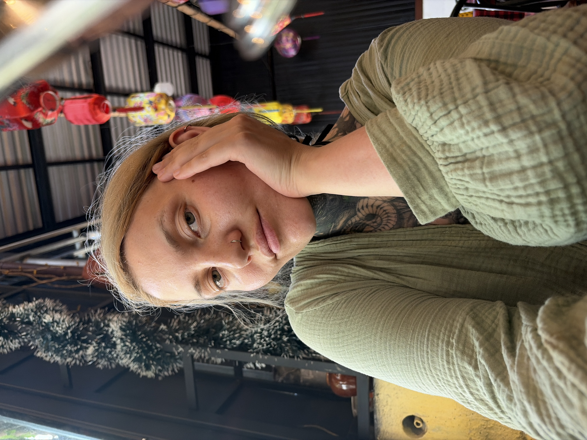 A young person, likely female, with blonde hair and light-colored eyes, is captured in a portrait-style shot. She has a septum nose ring and an earring in her right ear. Her left hand is gently placed on her temple or forehead, and she has visible dark tattoos on her neck/shoulder area and arm. She is wearing a light olive green, long-sleeved top made of a distinctly textured, crinkled or gauze-like fabric.
The setting appears to be an indoor or semi-outdoor casual establishment, such as a cafe, restaurant, or shop, consistent with the provided location context of Đà Nẵng, Vietnam. The background features several colorful, traditional-style lanterns, predominantly red, yellow, and purple, hanging from the ceiling. Below these, fuzzy garland-like decorations, possibly tinsel, are visible. The ceiling or upper wall structure is made of corrugated metal or slatted material. In the lower part of the frame, a dark surface, possibly a table, is visible, along with a yellow surface that has a dark, circular opening.
The person looks directly at the camera with a neutral to slightly contemplative expression, indicating a posed photograph. The lighting suggests it is daytime, likely with ambient or indirect light indoors. There is no visible text in the image.