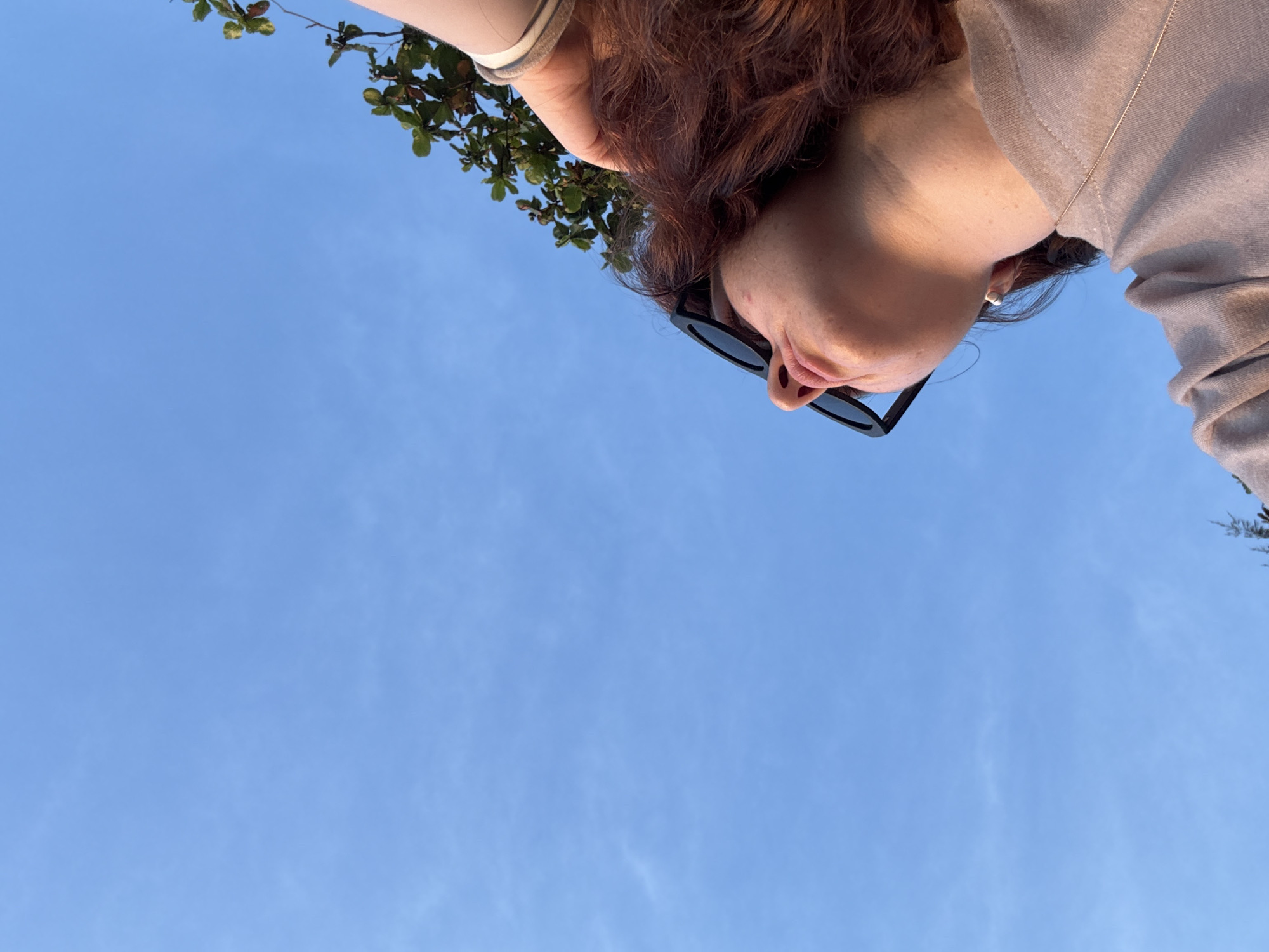 A low-angle, medium shot captures a person from the neck up, looking upwards against a bright, cloudless blue sky. The individual, positioned on the right side of the frame, has fair skin with freckles and auburn, wavy hair. They are wearing black sunglasses, a light-colored t-shirt, and a thin necklace. Their right hand is visible near their head, and a white wristband is present. A small branch with green leaves extends into the upper right corner of the frame. The lighting suggests it is daytime, possibly late afternoon, with warm light illuminating the person's face and neck. The background is a clear, expansive blue sky, indicating an outdoor setting. The specific location of Karon, Thailand is not discernible from the visual information present in the image.
