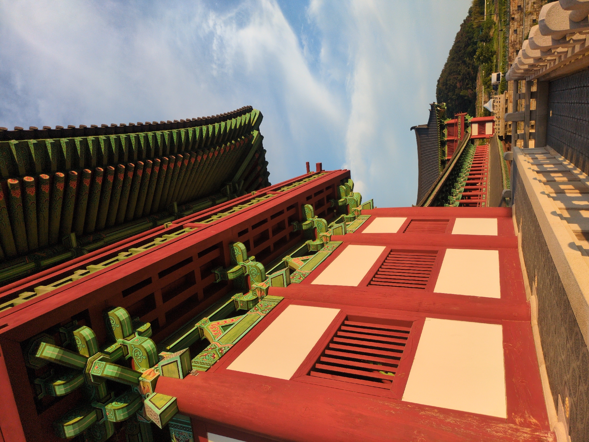 The image shows an upward-angled view of the traditional architecture of a building in Gyeongju-si, South Korea. The foreground features the red wooden framework of the building, adorned with green and red decorative elements. Above and to the left, the green tiled roof of a traditional Korean building is visible against a partly cloudy blue sky. To the right, the image shows the side of another building and a stone balustrade. A hill with trees is visible in the background.