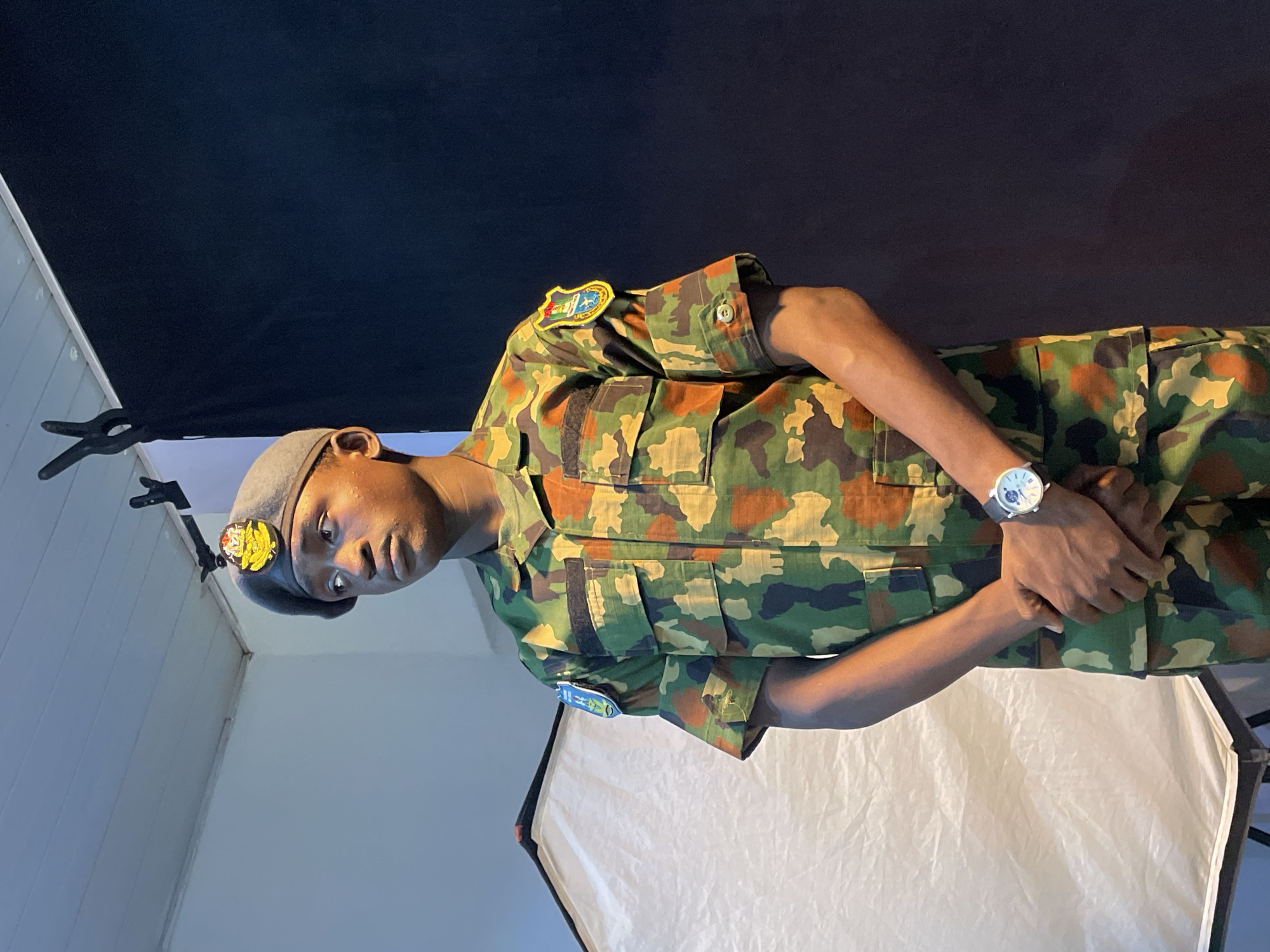 A man in military camouflage uniform stands in what appears to be a studio setting. He wears a beret with an emblem, and a watch on his left wrist. Behind him are studio lighting equipment, including a softbox and black backdrop. There are no details identifying a specific location in Barakin Akawo, Nigeria. The scene depicts a portrait setup, possibly for a professional photograph or identification.