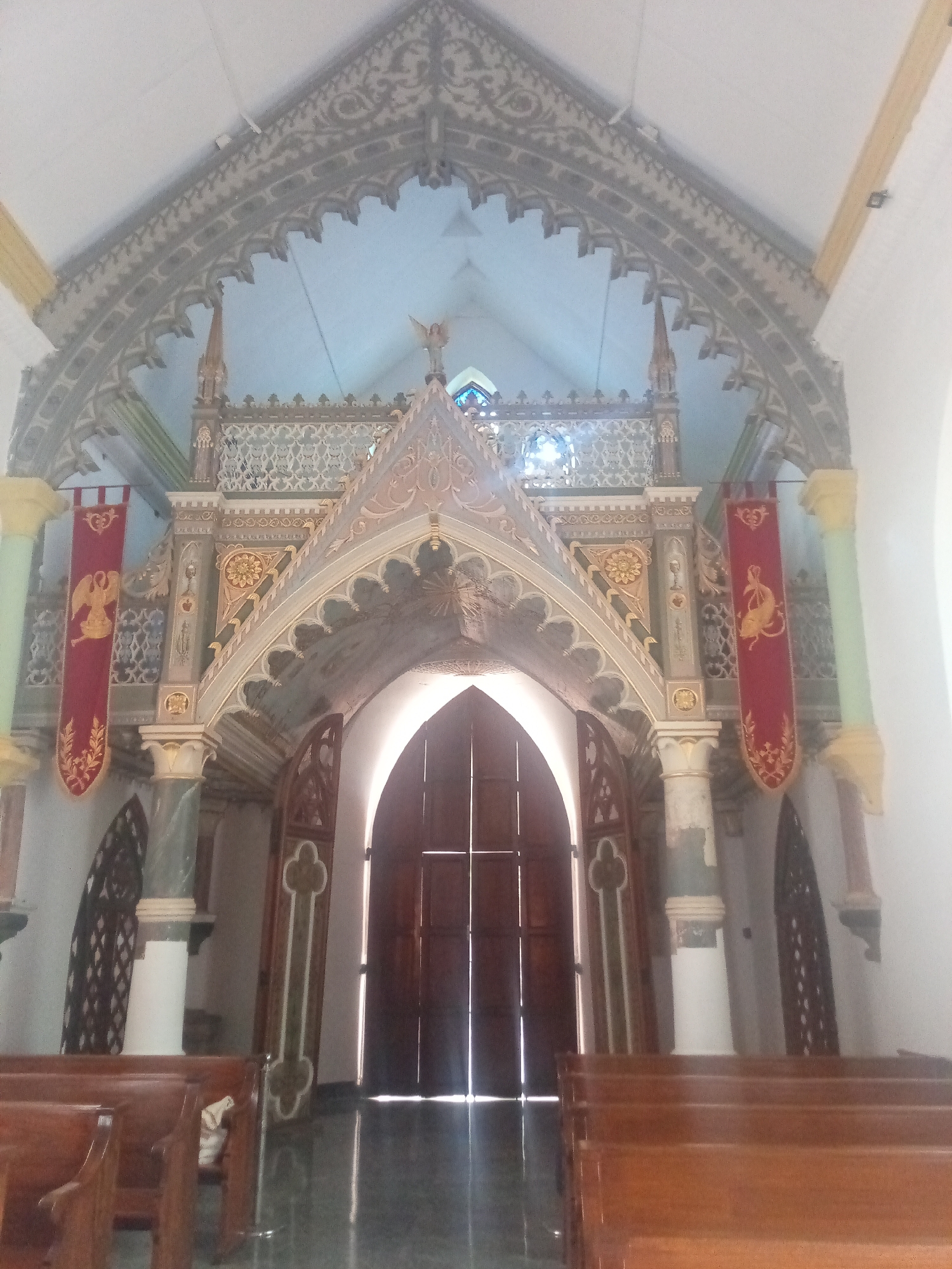 The image shows an interior view of a church in El Valle del Espíritu Santo, Venezuela. The architectural style is Gothic Revival, indicated by pointed arches and intricate decorative elements. The primary focus is the entrance to the sanctuary, framed by a highly ornate structure with columns, arches, and decorative screens. Two red banners with gold-colored emblems hang on either side of the entrance. Light streams through the closed wooden doors, illuminating the polished floor. In the foreground are rows of wooden pews.