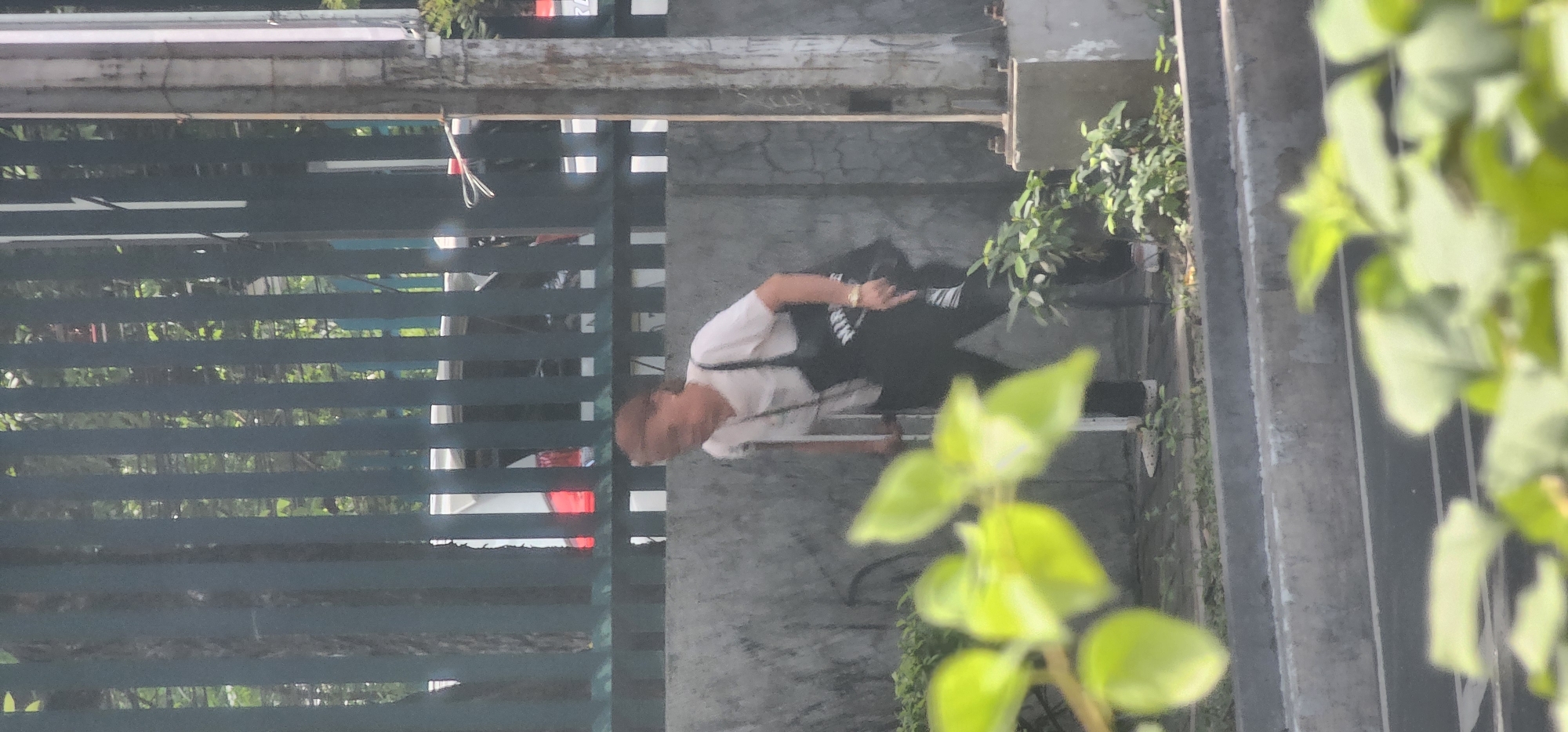 An elderly person with short, reddish-brown hair is walking with the assistance of a crutch, which is being used on their right side. They are wearing a white t-shirt and dark pants, and a dark bag is slung over their left shoulder. The person is on a concrete sidewalk, with a low concrete wall to their right and a dark metal fence behind them. Beyond the fence, a white vehicle is partially visible, with a red taillight showing. Green foliage is in the foreground, obscuring part of the scene. The location is identified as Quezon City, Philippines.