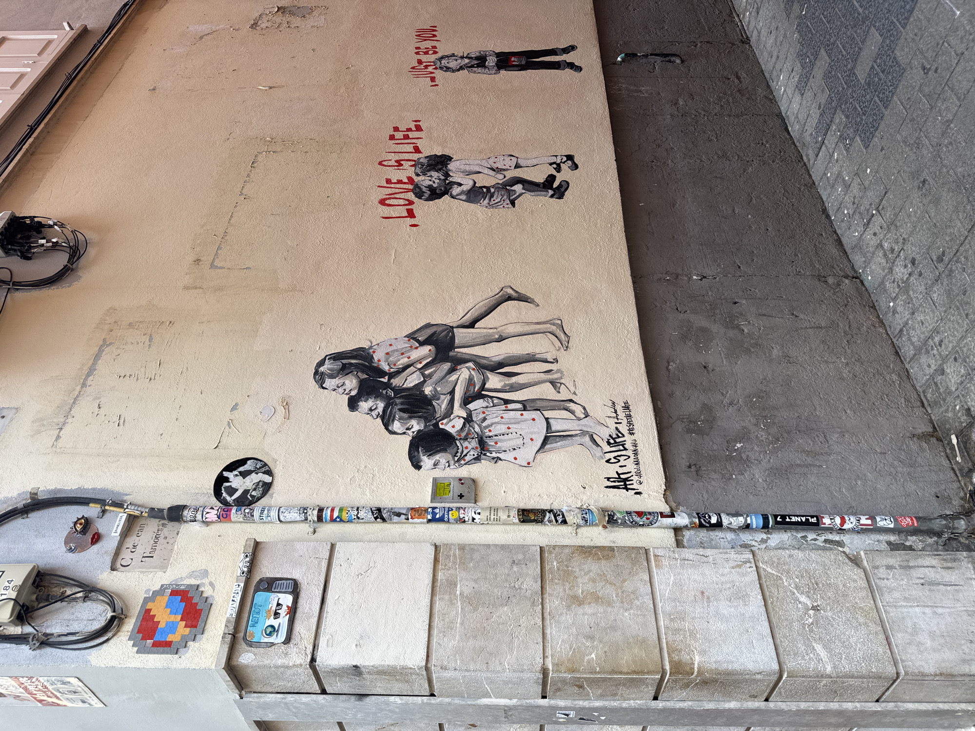 This image depicts a narrow street scene in Palma de Mallorca, Spain, featuring a building corner with graffiti and street art. The wall on the right is painted a light beige, with a section of rough, dark grey concrete at the base. The wall on the left is constructed of large, light-colored stone blocks. A utility pole, covered in numerous stickers, runs vertically along the corner.
On the beige wall, there are several pieces of street art. A large black and white mural shows a group of children, with a smaller mural of two children kissing and a single child holding a bucket. Red text is painted above the murals, reading ".LOVE IS LIFE." and ".JUST BE YOU.". Near the top left of the wall, a small mosaic featuring a pixelated red, yellow, and blue circle is adhered, along with a small, realistic eye with a red iris. A miniature television screen with a cartoon depiction of a dog wearing sunglasses is also present.
The ground is paved with cobblestones, a mix of grey and darker patterned stones. The overall impression is one of an urban environment with artistic expression integrated into its infrastructure.