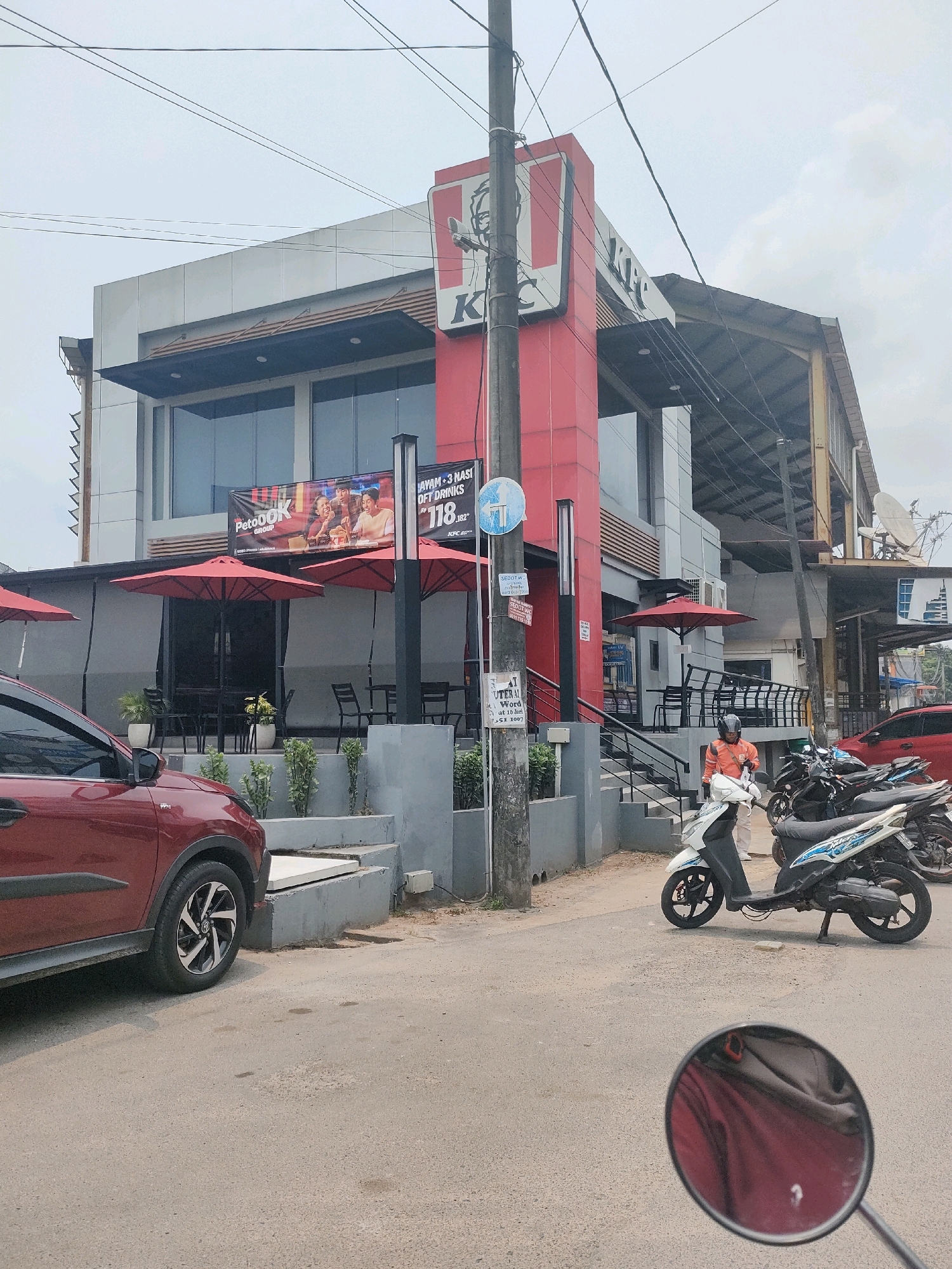 This image shows a KFC restaurant in Batam, Indonesia, with a red car parked on the left and several motorcycles on the right. The KFC building is modern, with large windows and a prominent red and white logo. Outside the restaurant, there are red umbrellas covering outdoor seating areas. A man wearing an orange jacket is standing near the motorcycles. The street is paved, and there are power lines visible overhead. The sky is overcast. The image appears to have been taken from a moving vehicle, as indicated by the reflection in a rearview mirror at the bottom of the frame.