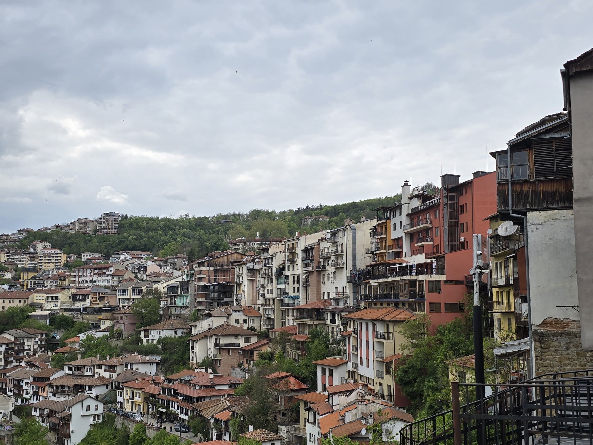 The image depicts a densely built urban landscape, likely a hillside town. The buildings are a mix of residential and commercial structures, characterized by a variety of architectural styles and colors. Red tile roofs are prominent, particularly in the foreground. In the background, a green, tree-covered hill rises above the town. The sky is overcast, with a layer of gray clouds. Some structures are equipped with antennas and satellite dishes. The image is taken from an elevated viewpoint, providing a panoramic view of the town. There is no observable presence of people or active human interaction. The location is an urban area, characterized by dense construction and varied architectural styles.