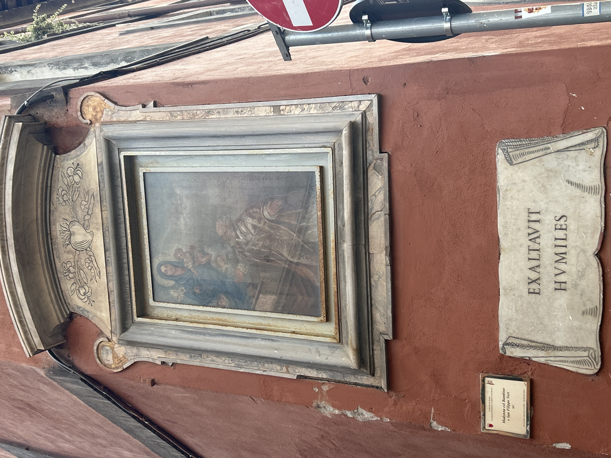 The image shows an exterior corner of a building in Rome, Italy. The wall is painted a reddish-brown. Mounted on the wall is a framed painting, depicting the Virgin Mary holding the infant Jesus, with a figure of a man kneeling before them. The painting is ornate, with a carved frame. Below the painting, there is a marble plaque with text in Latin, which reads "EXALTAVIT SVMILLES". To the left of the plaque, there is a smaller plaque with Italian text, identifying the painting as "Madonna col Bambino e San Filippo Neri". To the right of the painting, a street sign is visible. The scene appears to be in an urban setting.