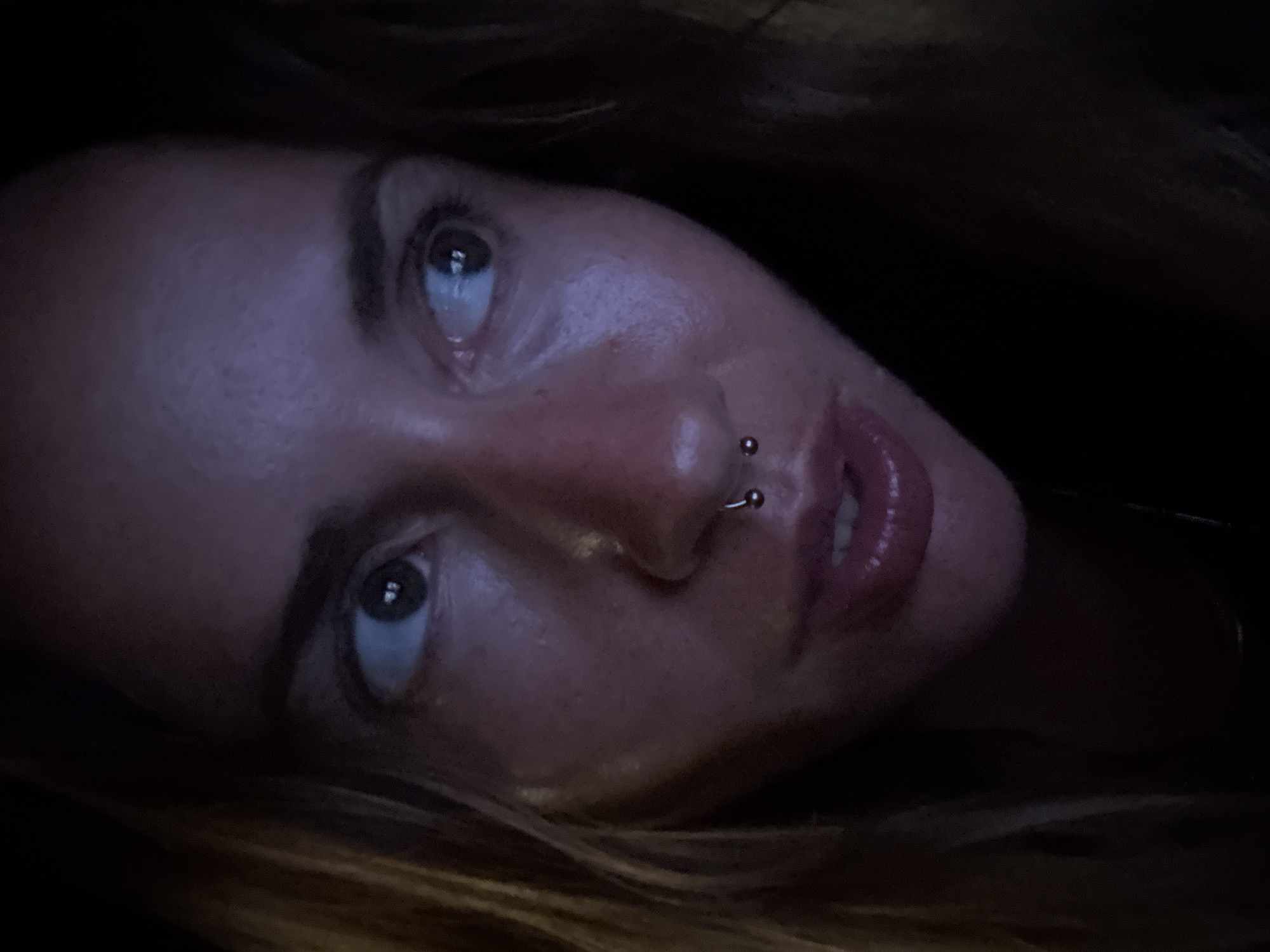 A close-up, head-and-shoulders portrait of a woman is captured in a dimly lit, indoor environment. The woman, whose face is turned to her right, gazes outward with wide eyes. Her hair, a medium brown, frames her face and falls loosely down her left side. She has a septum piercing with a small, silver horseshoe-shaped stud. Her lips are parted slightly, revealing her teeth, and appear to be glossed. The lighting casts a purplish hue on her skin, highlighting the texture and some minor blemishes. The background is dark, obscuring any details of the surrounding environment, which is presumed to be within the city of Unknown, Unknown.