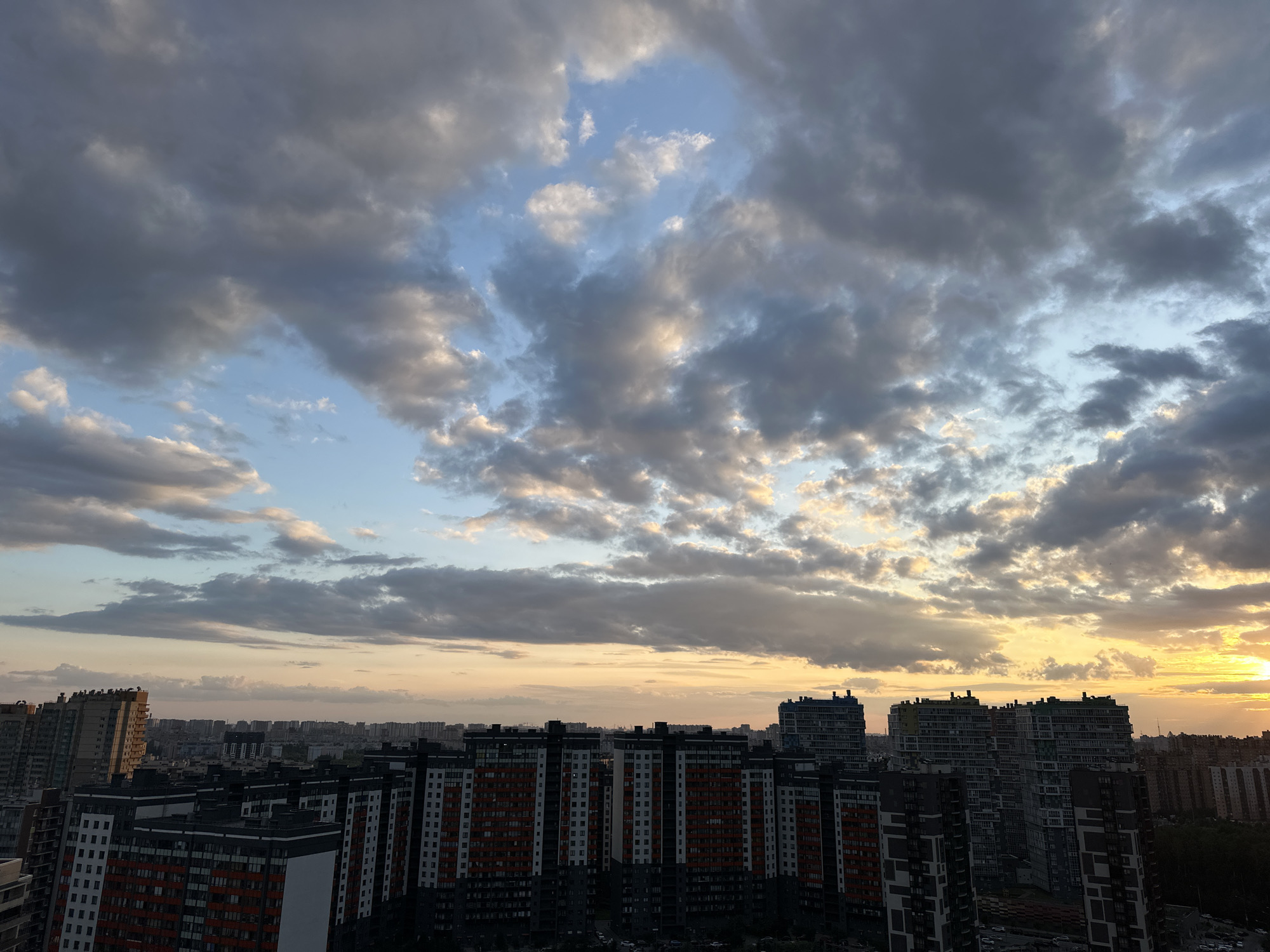 The image depicts a wide-angle aerial view of a dense urban landscape in Sinyavino, Russia, under a dramatic sunset sky. The foreground is dominated by rows of modern, multi-story residential buildings, characterized by their dark facades with accents of orange, white, and gray. These buildings are tall and appear to be densely packed. In the middle ground and background, more buildings of varying heights stretch towards the horizon, creating a sprawling cityscape. The sky is filled with a dynamic cloud formation; dark, heavy clouds are interspersed with patches of bright blue sky, and the setting sun casts a warm, golden glow on the horizon and illuminates the edges of some clouds. The overall impression is one of a vast, modern city at the end of the day, with nature's celestial display providing a striking backdrop.