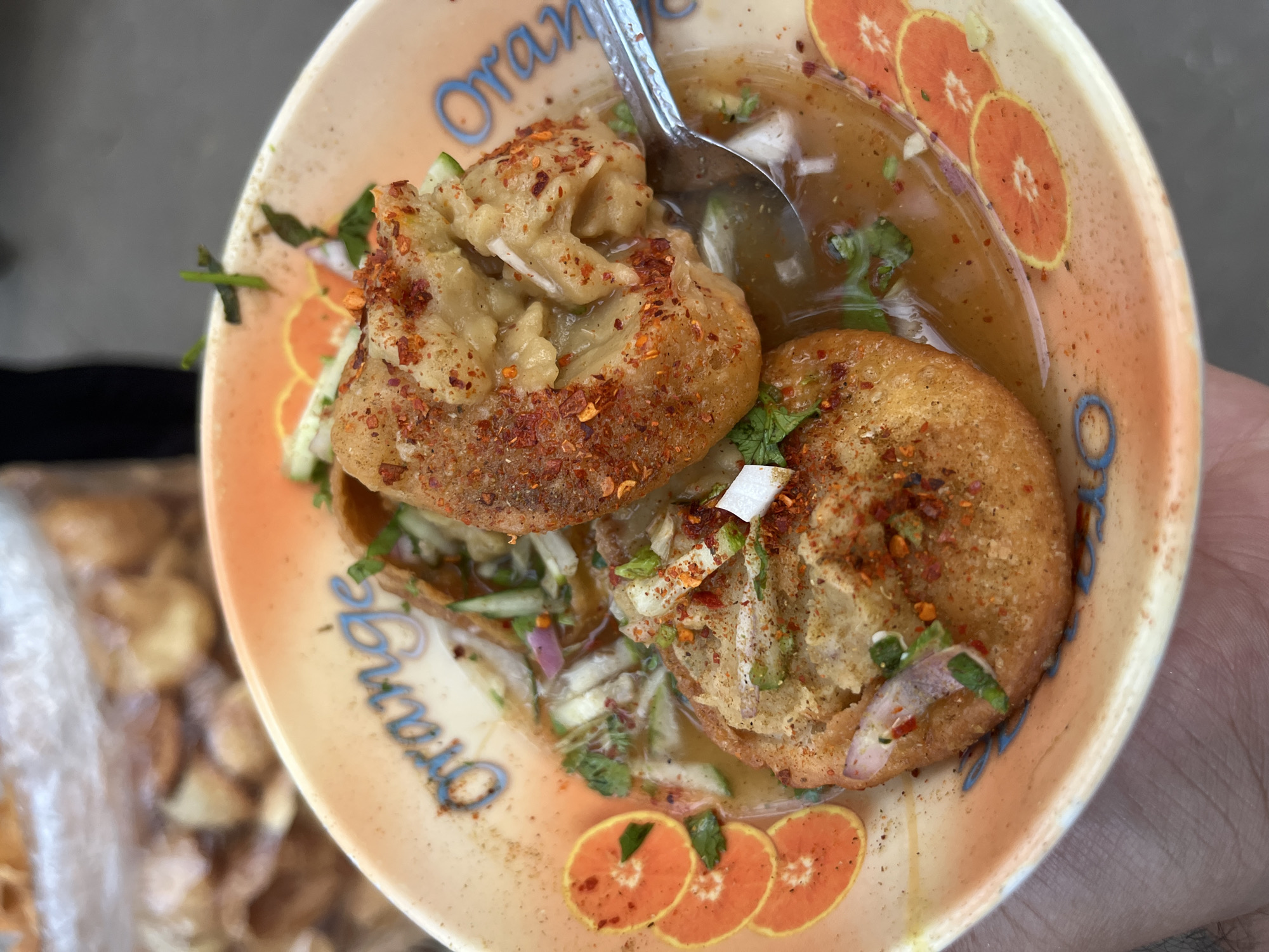 A close-up, high-angle shot captures a bowl held by a person's hand, containing a popular South Asian street food dish, likely Fuchka or Golgappe, as commonly found in Dhaka, Bangladesh.
The bowl holds two prominent, golden-brown fried shells, which are generously filled with a mashed, yellowish mixture, appearing to be spiced potatoes and chickpeas. These filled shells are partially submerged in a tangy, reddish-brown liquid or broth, known as 'pani'. The dish is vibrantly garnished with fresh toppings including finely chopped green herbs (cilantro), white and purple onion pieces, and green cucumber slivers. A significant amount of red chili flakes and other spices are liberally sprinkled over the entire dish, enhancing its visual appeal and suggesting a spicy flavor profile. A metal spoon is visible, partially immersed in the liquid, indicating that the dish is ready for consumption.
The serving bowl is light orange and decorated with several stylized orange slices around its inner rim. The word "Orange" is prominently written in a flowing blue cursive font on the interior side of the bowl. In the blurred background, a translucent plastic bag contains several light-colored, irregularly shaped items, possibly additional fried components or ingredients related to the meal. The scene evokes a casual, outdoor dining experience, characteristic of street food culture.