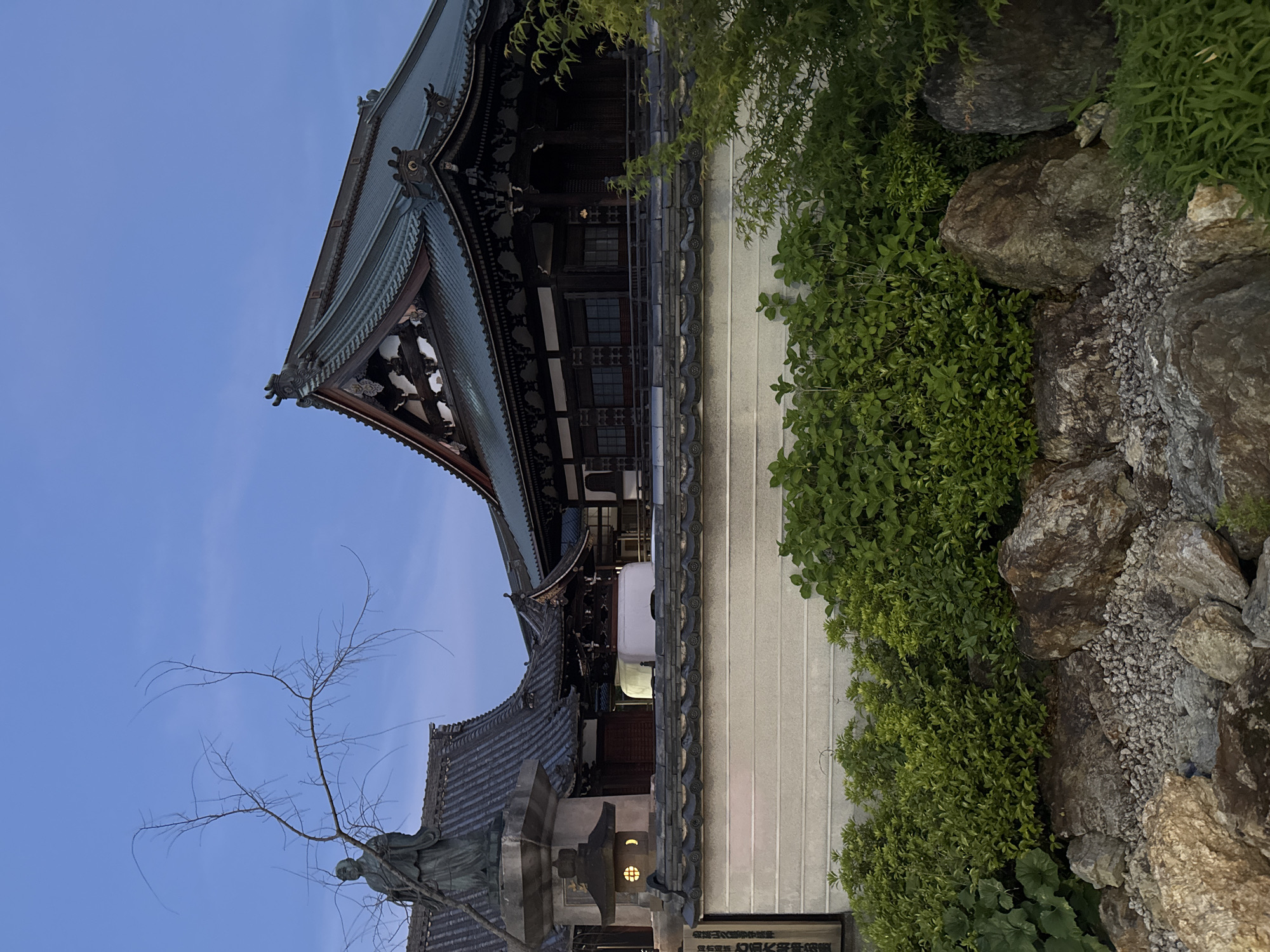 The image shows a temple in Kyoto, Japan, at dusk. The temple has a tiered roof with intricate eaves and ornamentation. Adjacent to the temple, a white wall stretches along the scene, lined with lush greenery and interspersed with rocks. In the foreground, a stone lantern stands near a statue, silhouetted against the sky. The sky transitions from a deeper hue at the top to a lighter shade at the bottom, suggesting the onset of night.