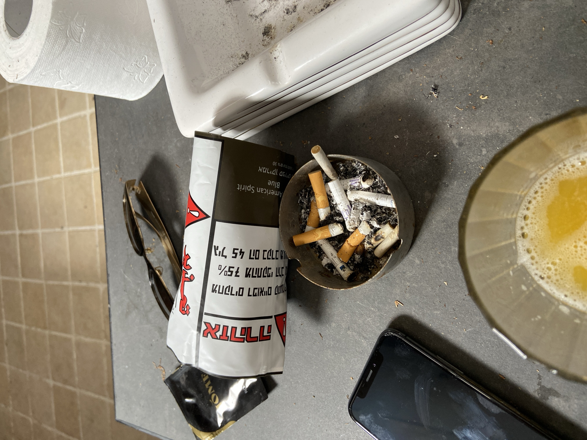 The image depicts a cluttered bathroom counter. A roll of toilet paper is adjacent to a dirty white porcelain sink. Beside the sink is an ashtray filled with cigarette butts, next to an opened pack of "Globes" cigarettes with Hebrew text and an "American Spirit Blue" pack peeking out behind it. A pair of glasses and a black smartphone also rest on the counter. Next to them is a glass filled with a yellowish liquid, possibly beer. The scene suggests a casual, perhaps unkempt, environment. There is no indication of Tel Aviv, Israel other than the Hebrew text on the pack of "Globes" cigarettes.