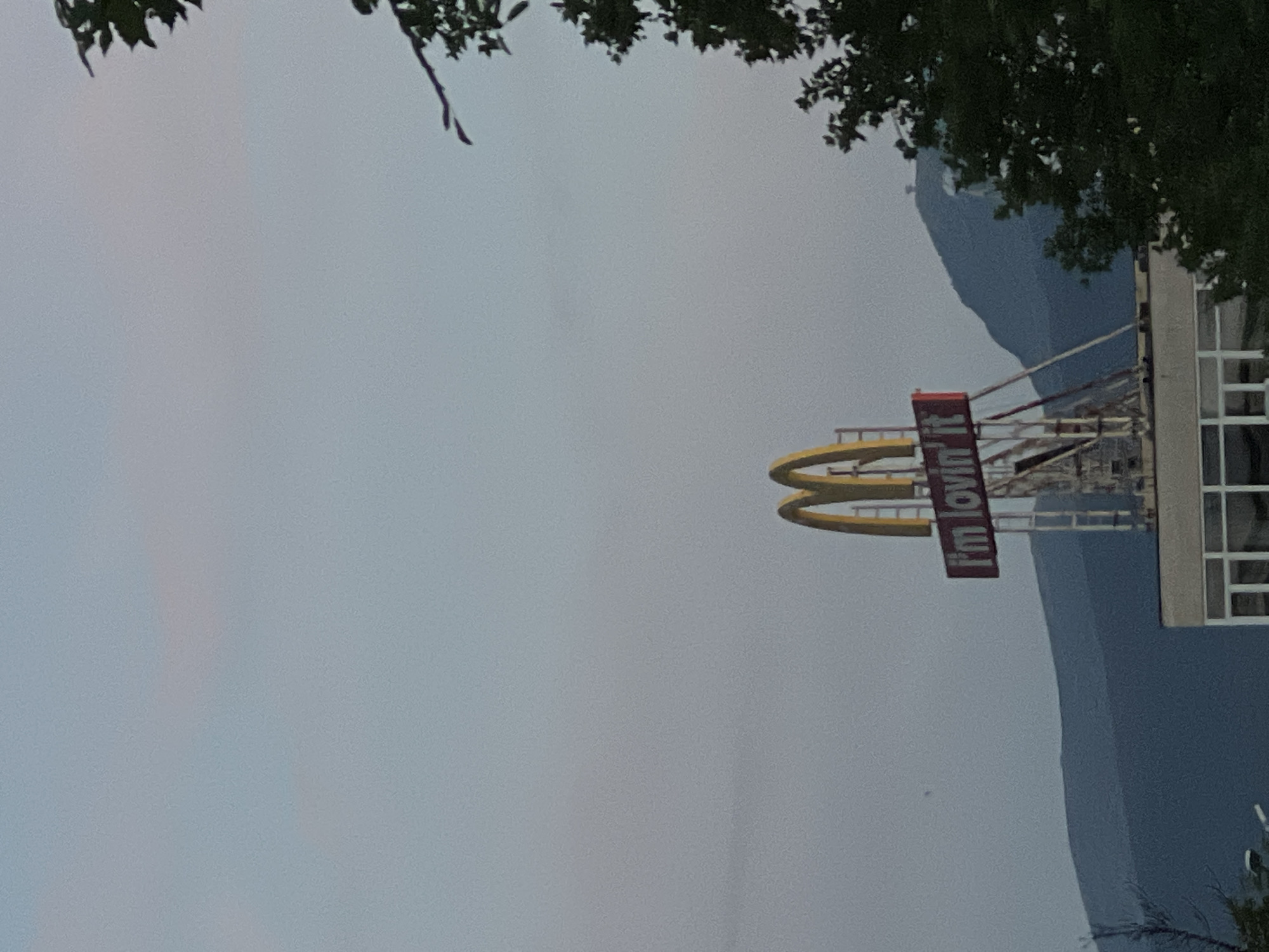The image shows a close-up of a McDonald's sign with the slogan "i'm lovin' it" against a muted, overcast sky. The iconic golden arches of the McDonald's logo are visible above the slogan. The sign appears to be mounted on a tall structure, possibly scaffolding, suggesting maintenance or construction work is underway. In the background, rolling hills or mountains are visible, shrouded in a hazy blue hue. A portion of a building with a white facade and prominent windows is visible at the bottom of the frame. Dark green foliage from trees frames the right side of the image. The overall lighting suggests it is either early morning or late evening. This McDonald's sign is located on the Sofia ring road, near the Lyulin highway interchange.