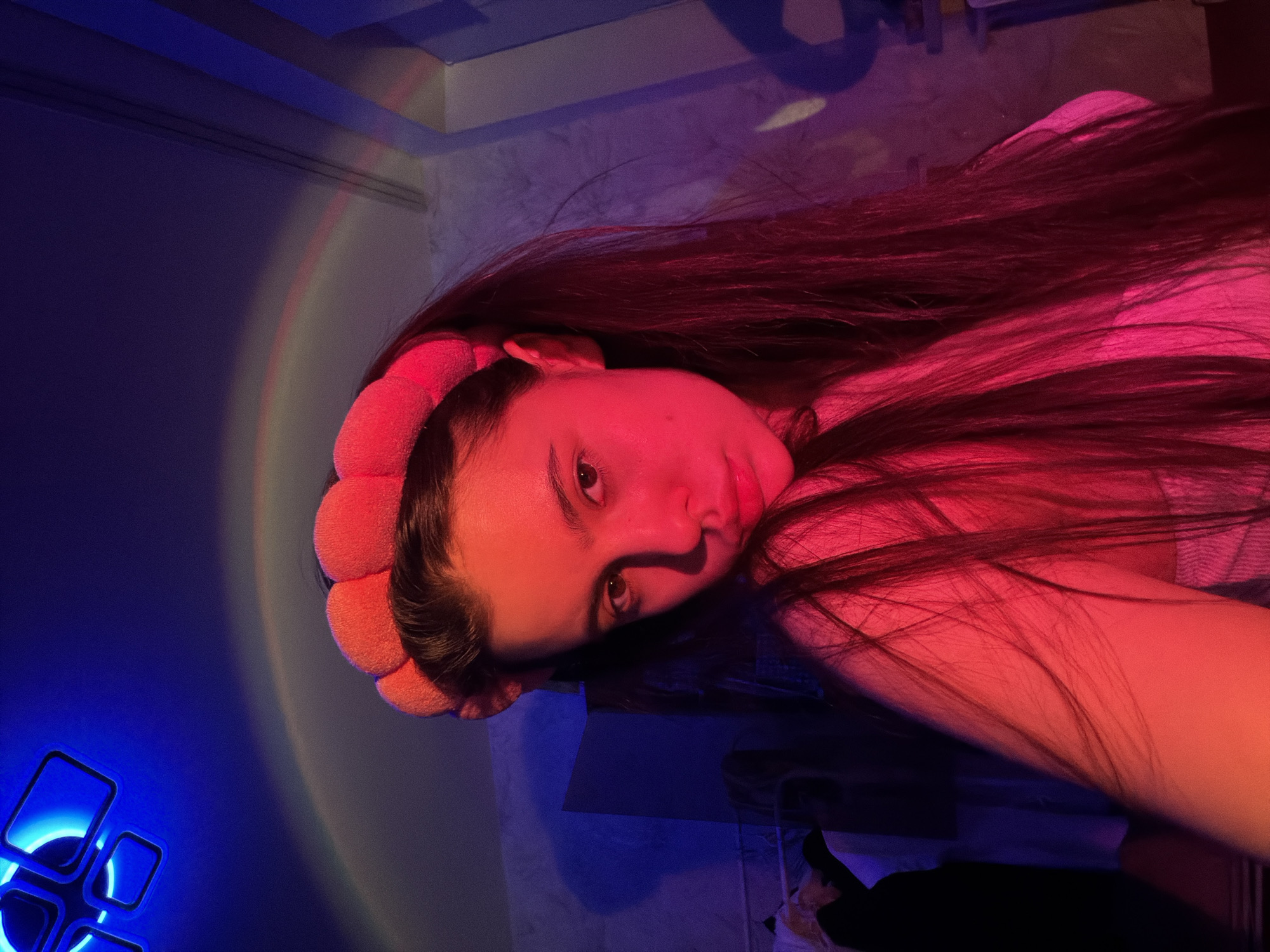 The image depicts a person lying down, likely indoors, in a room with ambient lighting featuring red and blue hues. The person has long, dark hair and is wearing a padded, orange-colored headband. There is a blue light source with geometric shapes on the wall to the left. A rainbow-colored projection also appears on the wall near the top left. The location is potentially Chaikovsky, Russia, though there are no specific details in the image itself to confirm this.