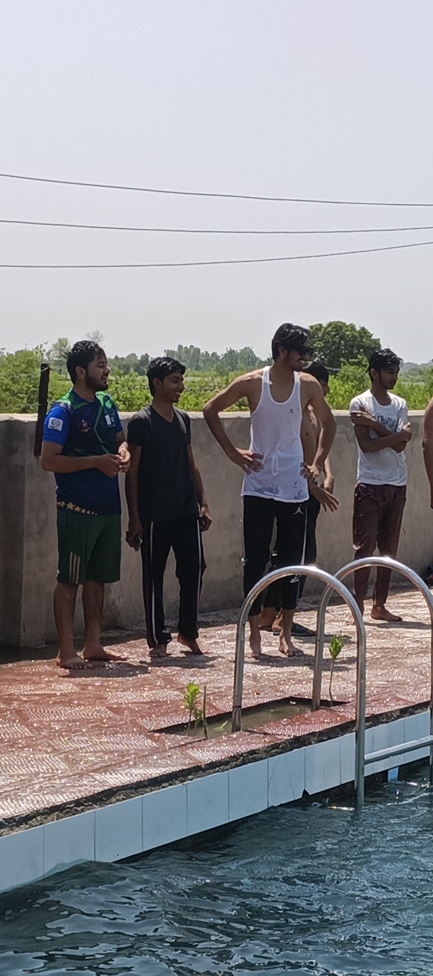 A group of five young men stand on the tiled edge of an outdoor swimming pool, located in Samundri, Pakistan, on a sunny day. The first man is wearing a blue and green Pakistan jersey with green shorts. The second man is in a black t-shirt and black pants. The third man wears a wet, white tank top and black pants. The fourth man is shirtless. The fifth man is wearing a white t-shirt and maroon pants. The pool has a metal ladder for access. Behind them is a low concrete wall and a rural landscape with greenery. Power lines are visible in the sky.