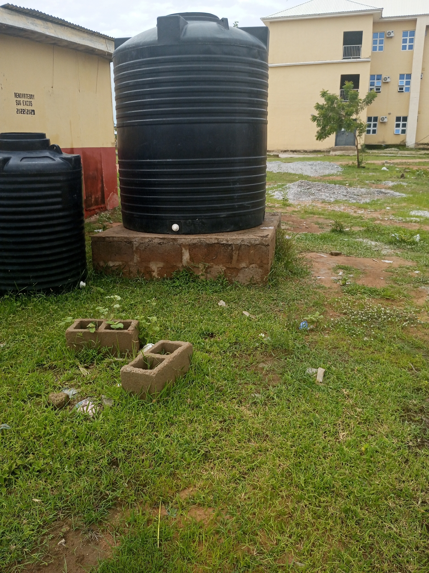 The image depicts an outdoor scene with two large black water tanks positioned on a grassy area. One tank, taller and more prominent, is elevated on a concrete block foundation. To its left, a smaller water tank sits directly on the ground. Behind the tanks, a building with a yellow facade and a corrugated metal roof is visible, featuring a red band along its lower section. Further in the background, another building with a cream-colored exterior and multiple windows is seen, with air conditioning units installed next to some of the windows. A small tree grows in the open space between the buildings.
The ground is covered in green grass, with patches of exposed soil and small piles of gravel. Several hollow concrete blocks, some containing small plants, are scattered on the grass in the foreground. The sky appears overcast, suggesting a cloudy day.
Text is visible on the yellow building to the left of the tanks. It reads:
"RENOVATED BY
SUG EXCOS
2022|2023"
The scene appears to be set in a relatively calm environment, likely a residential or institutional area, given the presence of multiple buildings. There is no indication of any specific activity or event taking place. The time of day is not discernible, but the overcast weather suggests it is daytime. The overall impression is one of utility infrastructure within an educational or residential compound.