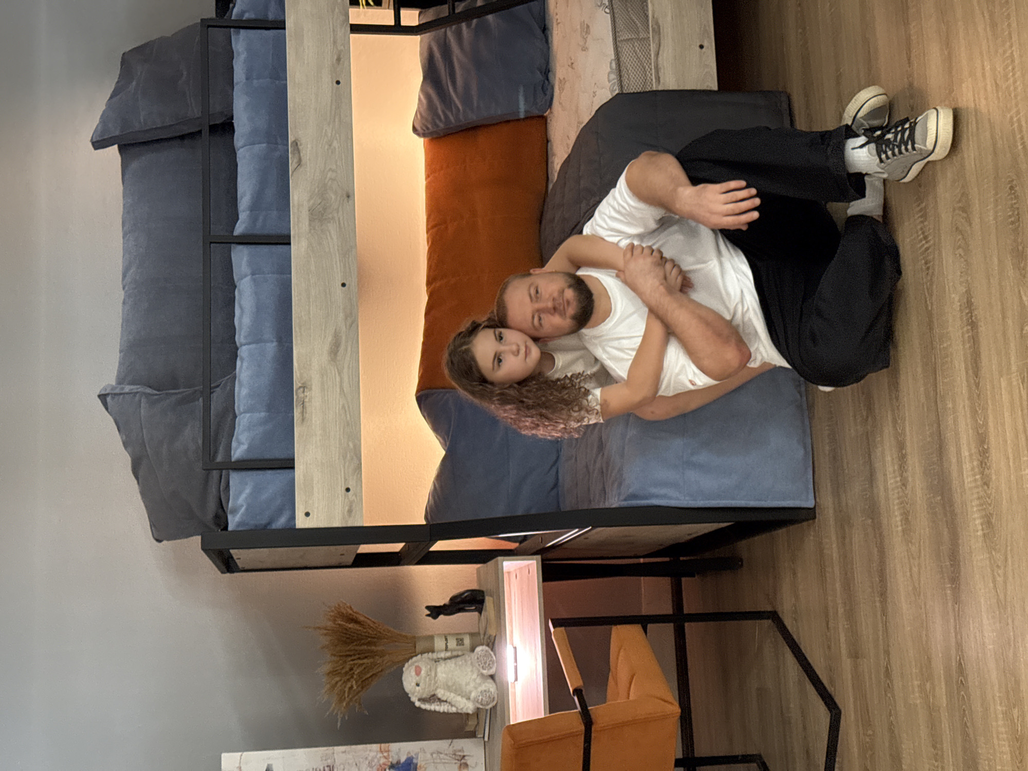 A medium shot captures a father and daughter posing in a bedroom. The father, with a beard and wearing a white t-shirt and black pants, sits on the wooden floor. His daughter, a young girl with curly brown hair, stands behind him, hugging him around the neck. She wears a white t-shirt. They are positioned in front of a bunk bed with blue and dark grey bedding. To the left of the bunk bed is a desk with an orange chair. On the desk are a stuffed rabbit, a black cat figurine, and a vase of dried grass. The room is lit by a warm, ambient light, and the floor is covered in wood-style laminate. The setting is likely a residential interior. No specific details indicating a location in Chaikovsky, Russia are visible.
